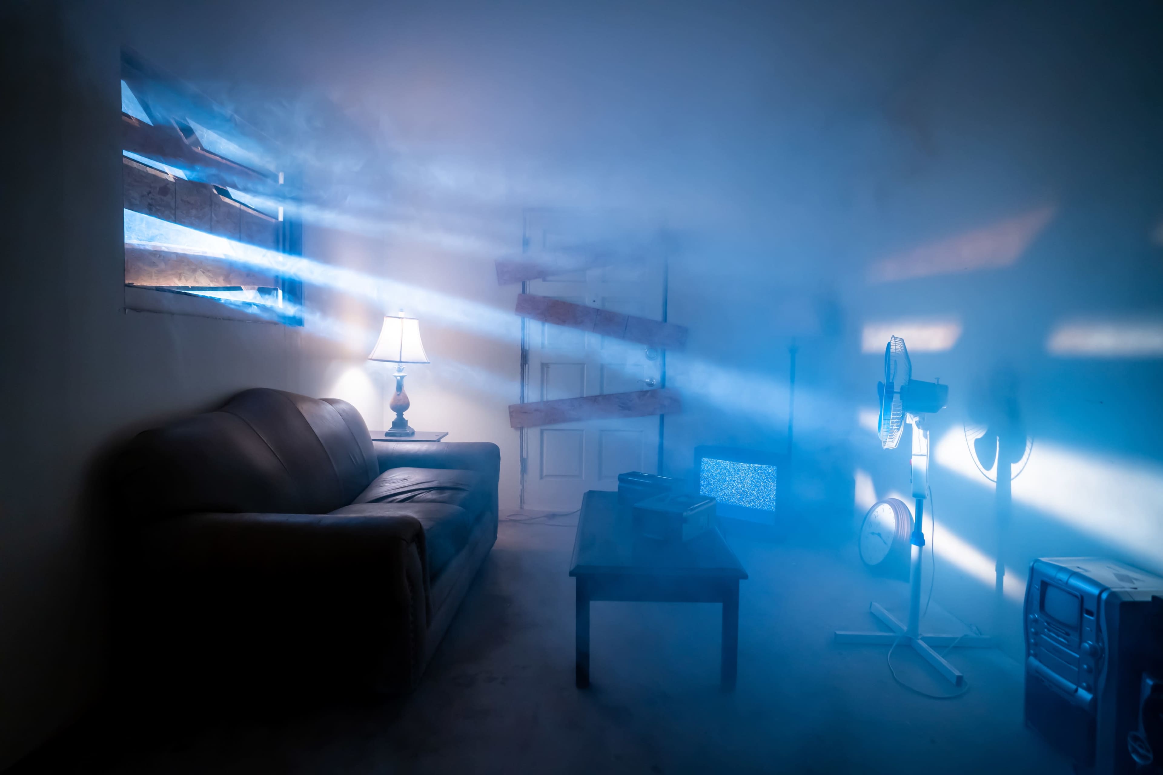 A dimly lit room features a couch, a table, a lamp, and a fan, with beams of light cutting through a mist and a boarded-up door.