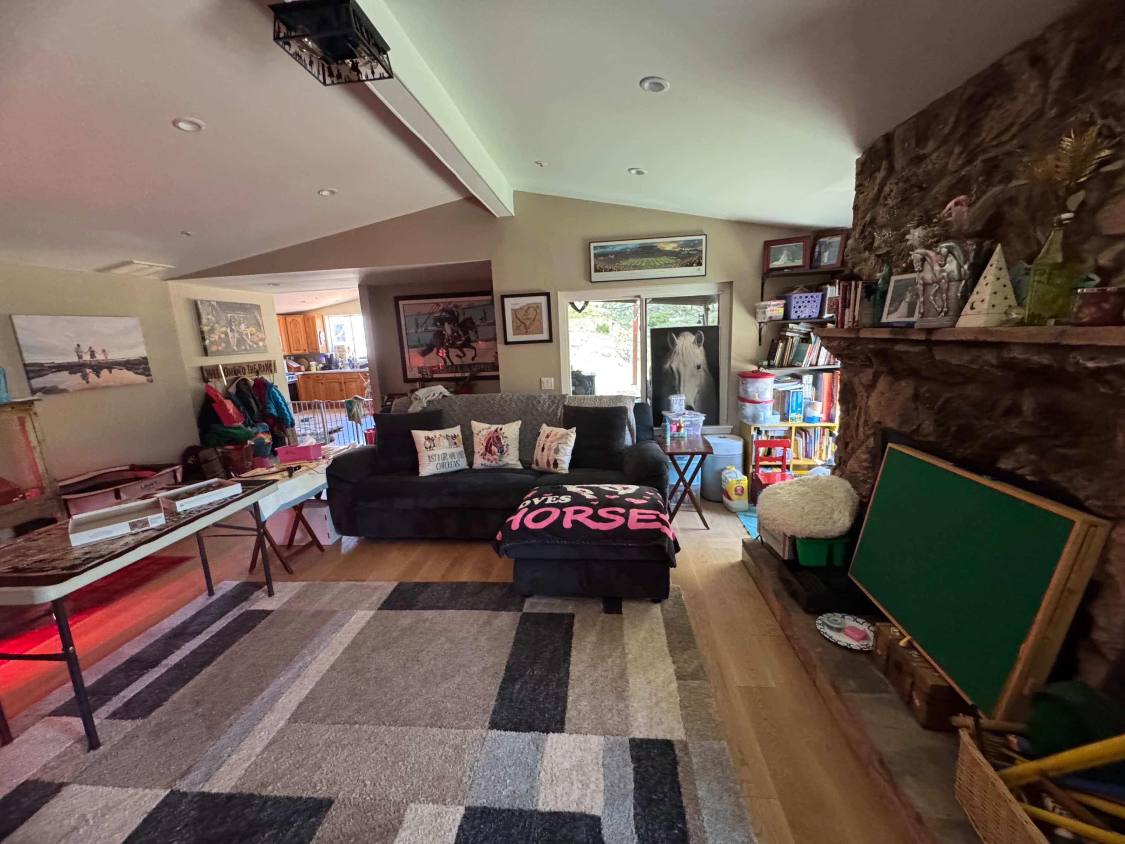 The image shows a cozy living room with a black couch, a large area rug, and a stone wall, featuring various decorations and a view of a kitchen area in the background.