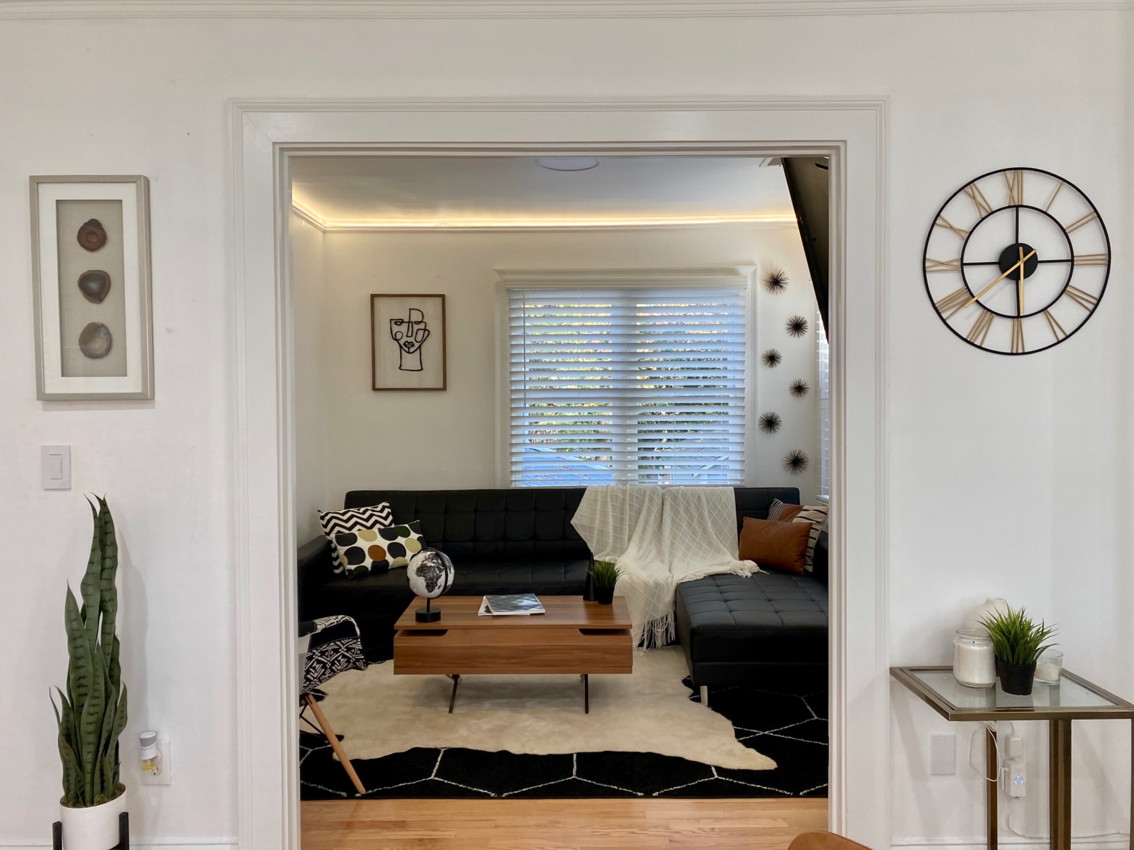 A living room features a dark sofa arrangement, a wooden coffee table, and decorative elements including a plant and a wall clock.
