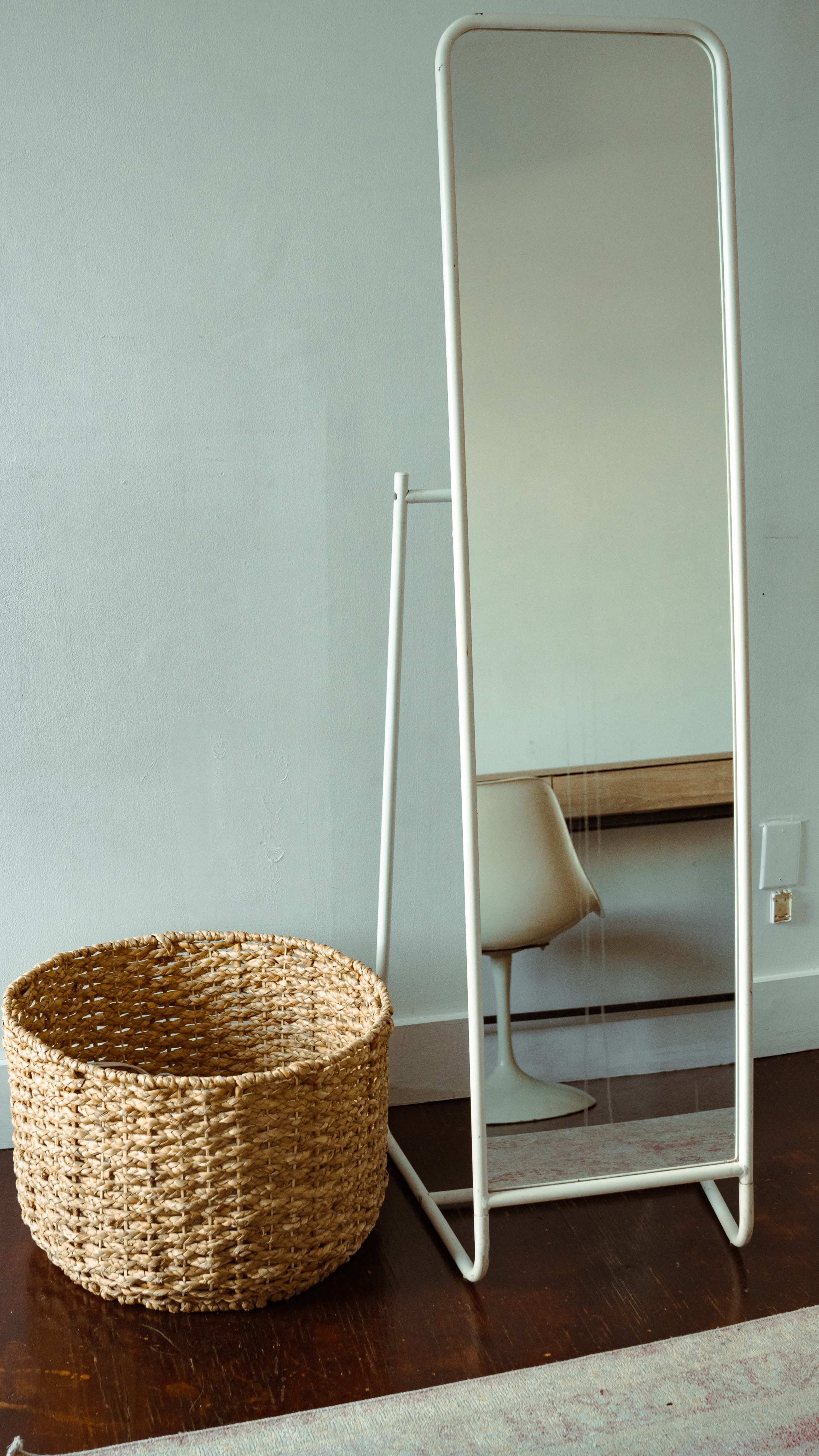 A tall, standing mirror with a white frame is positioned next to a woven, round basket on a wooden floor.