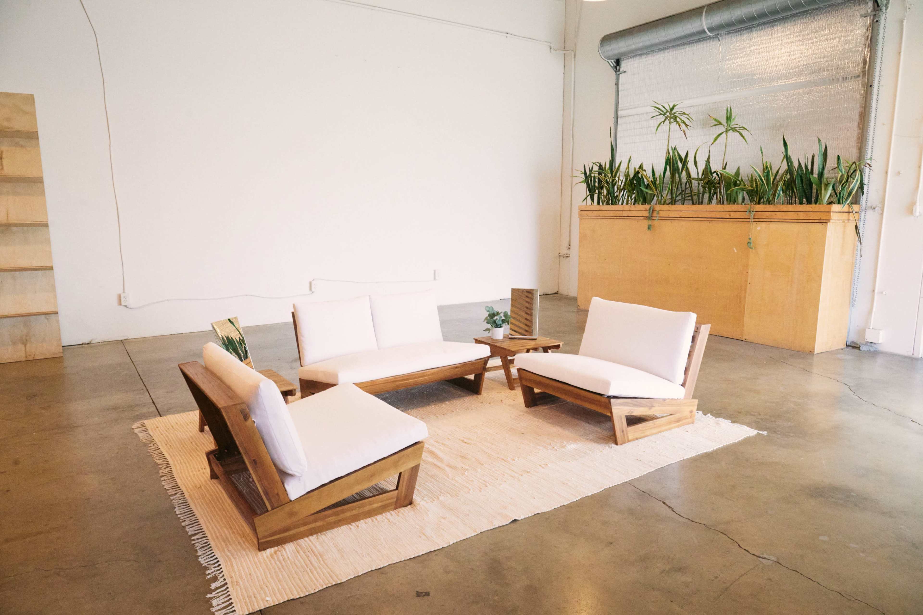 A minimalist seating arrangement with four wooden chairs and a low table on a beige rug in a spacious room with concrete flooring and potted plants.