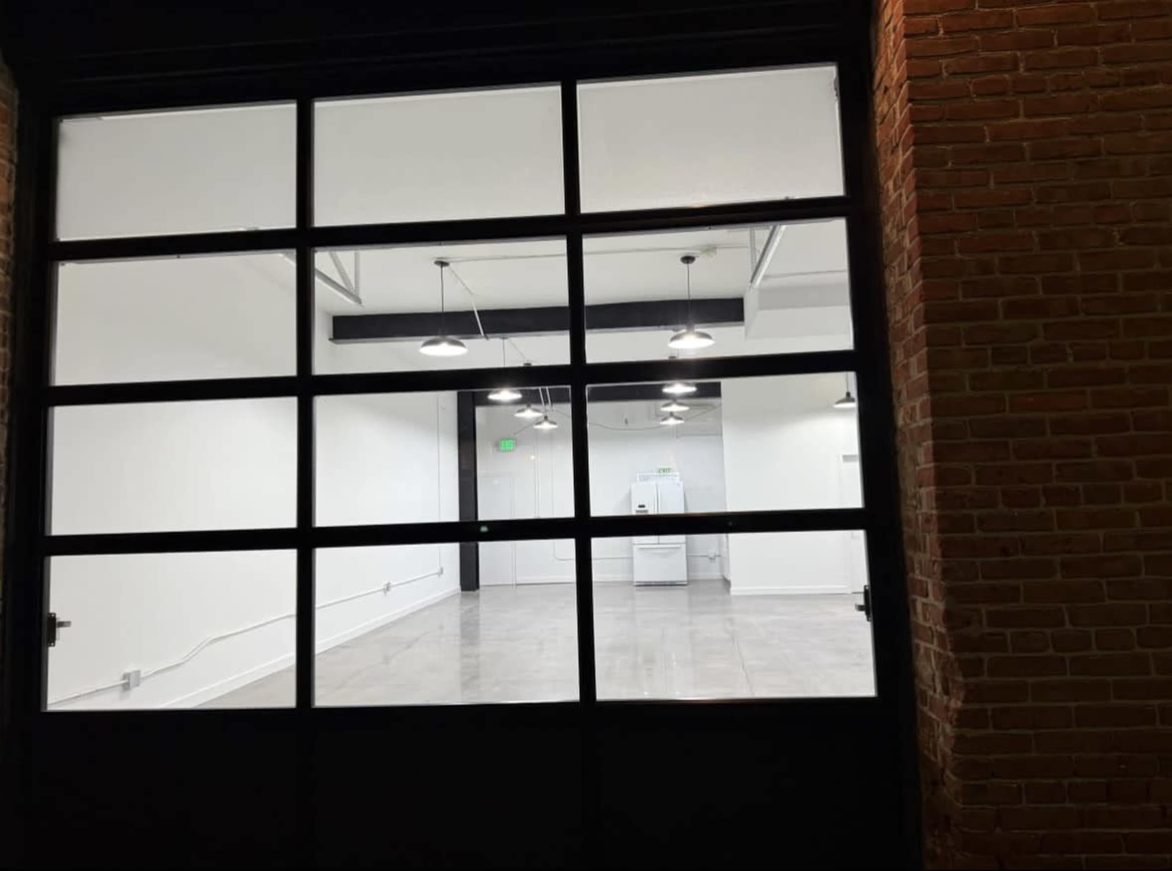The image shows a large, empty room with white walls and industrial-style lighting, viewed through a window with multiple panes.