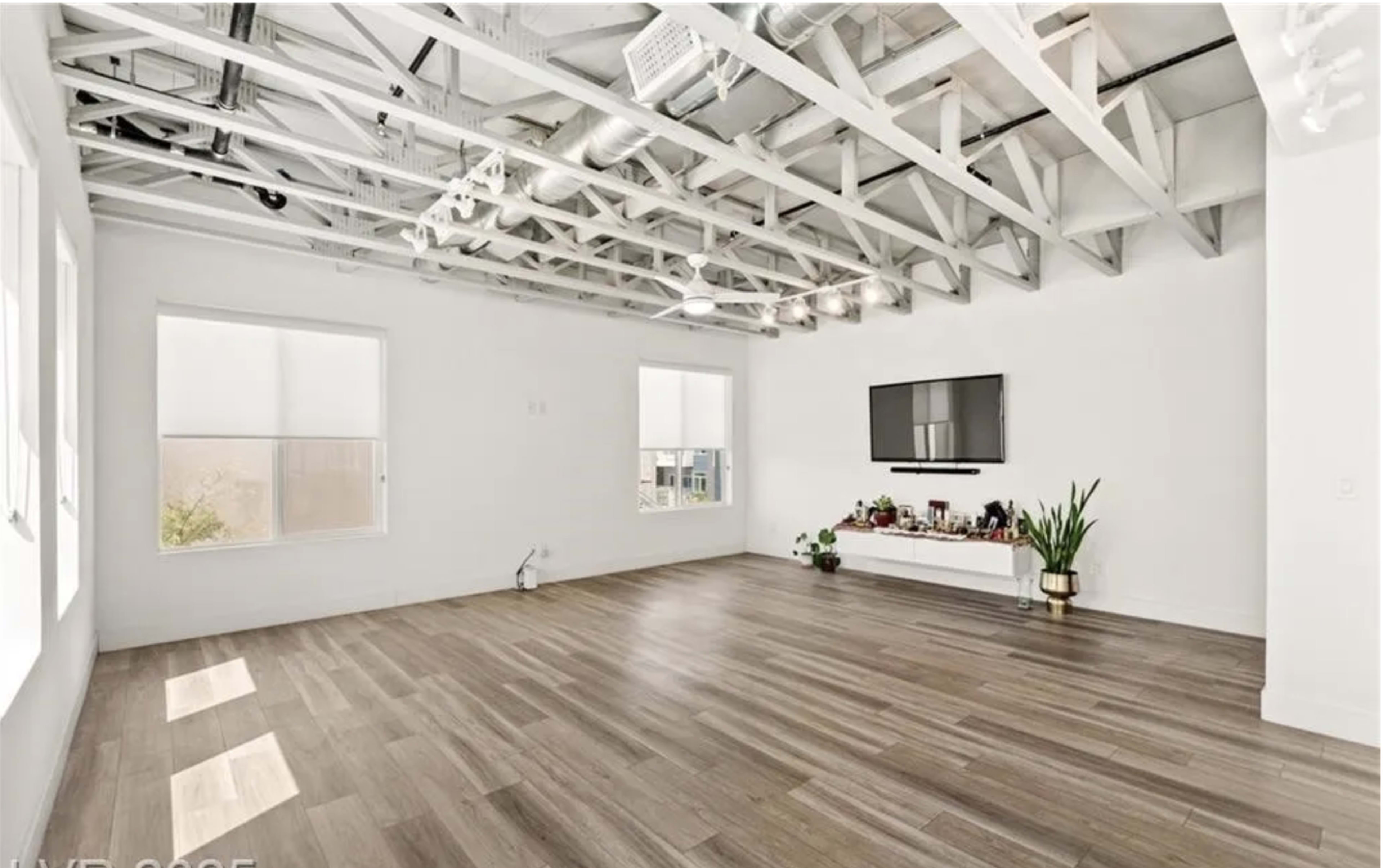 The image shows a spacious, empty room with wooden flooring, large windows, and a mounted television on a white wall, complemented by a small shelf featuring decorative items and a potted plant.