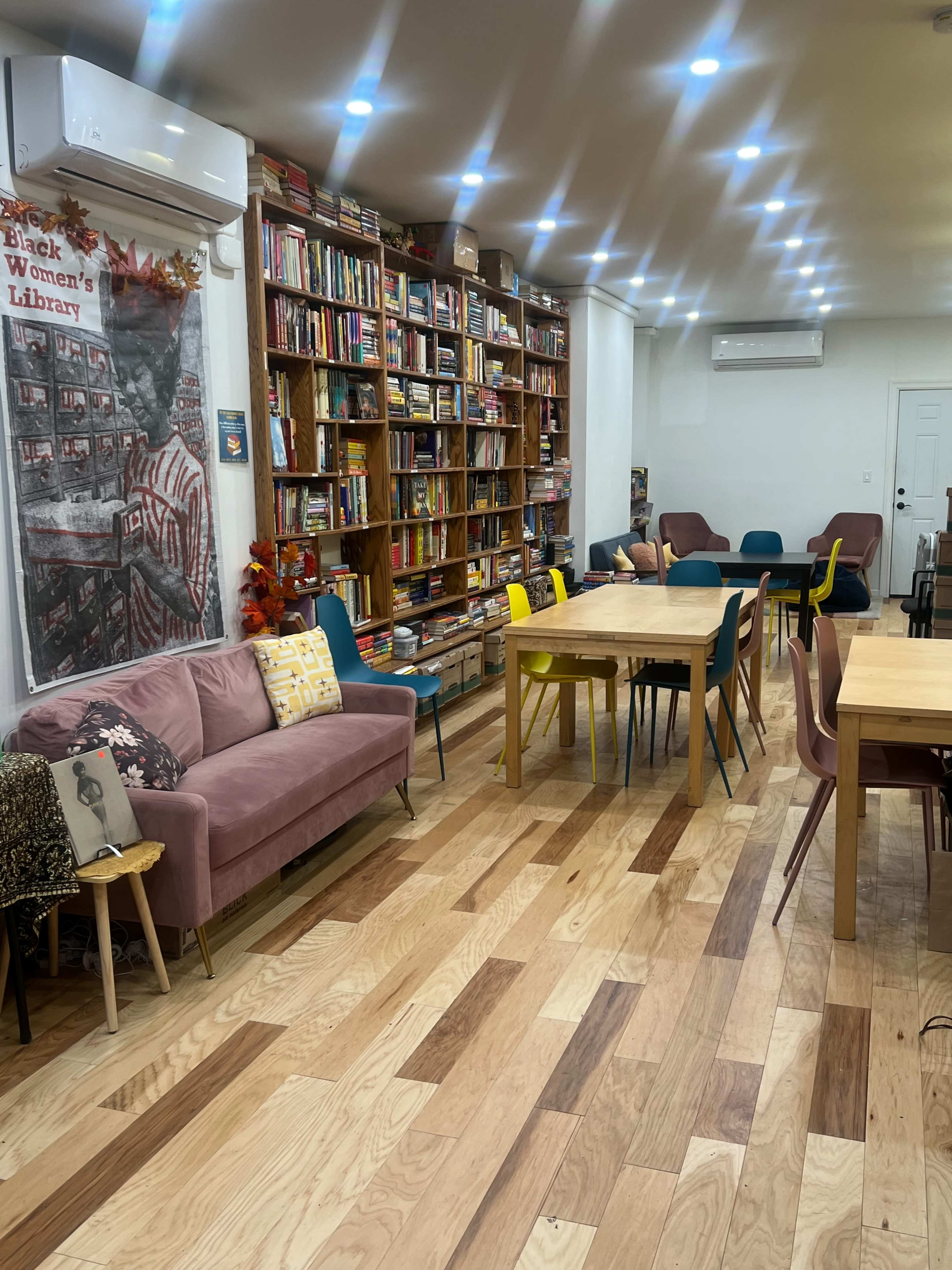 A cozy reading room with a bookshelf filled with books, several tables, and colorful chairs arranged around a wooden floor.