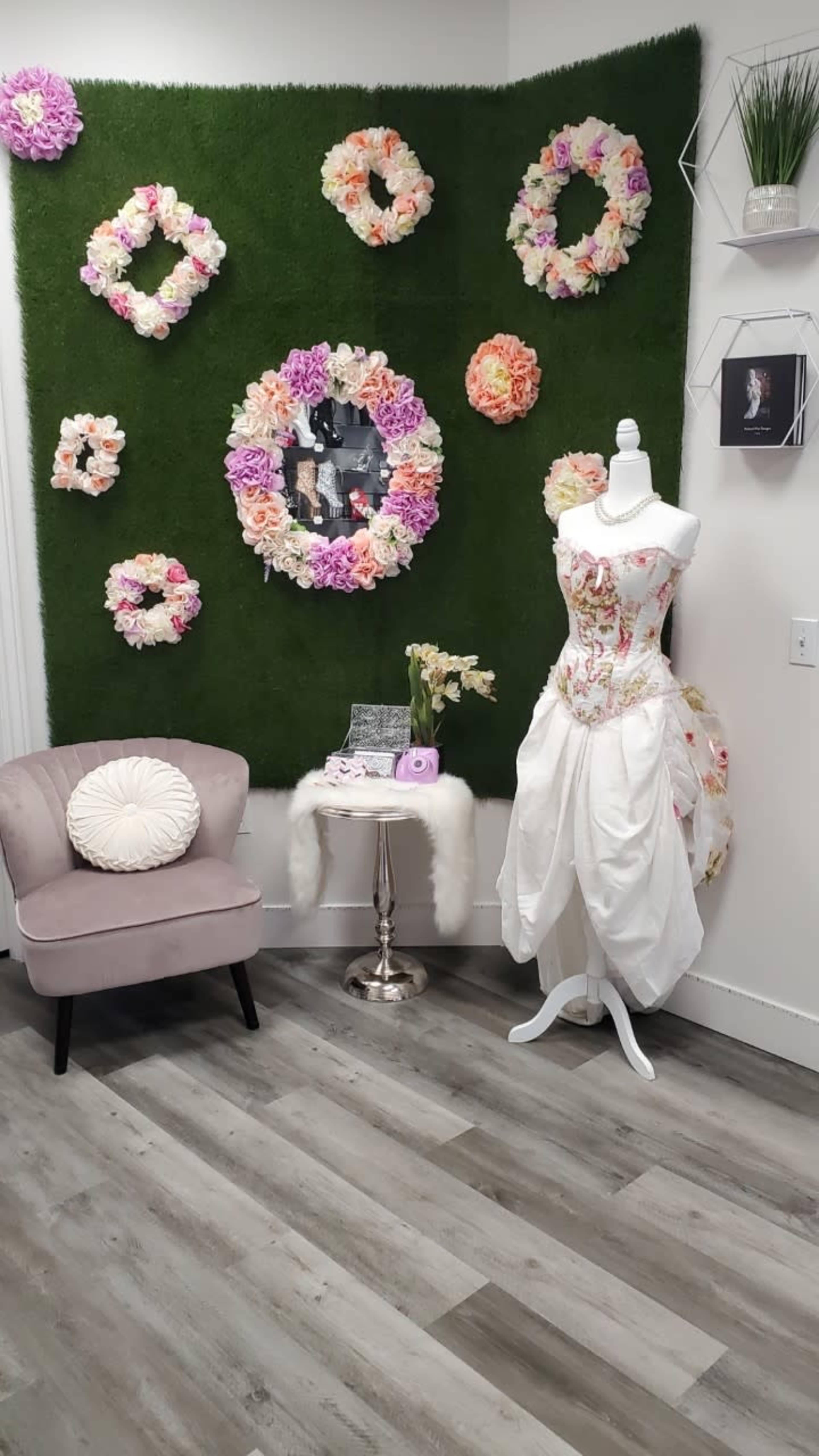 A dress form displays an intricately designed wedding dress next to a floral backdrop and a small table in a boutique setting.