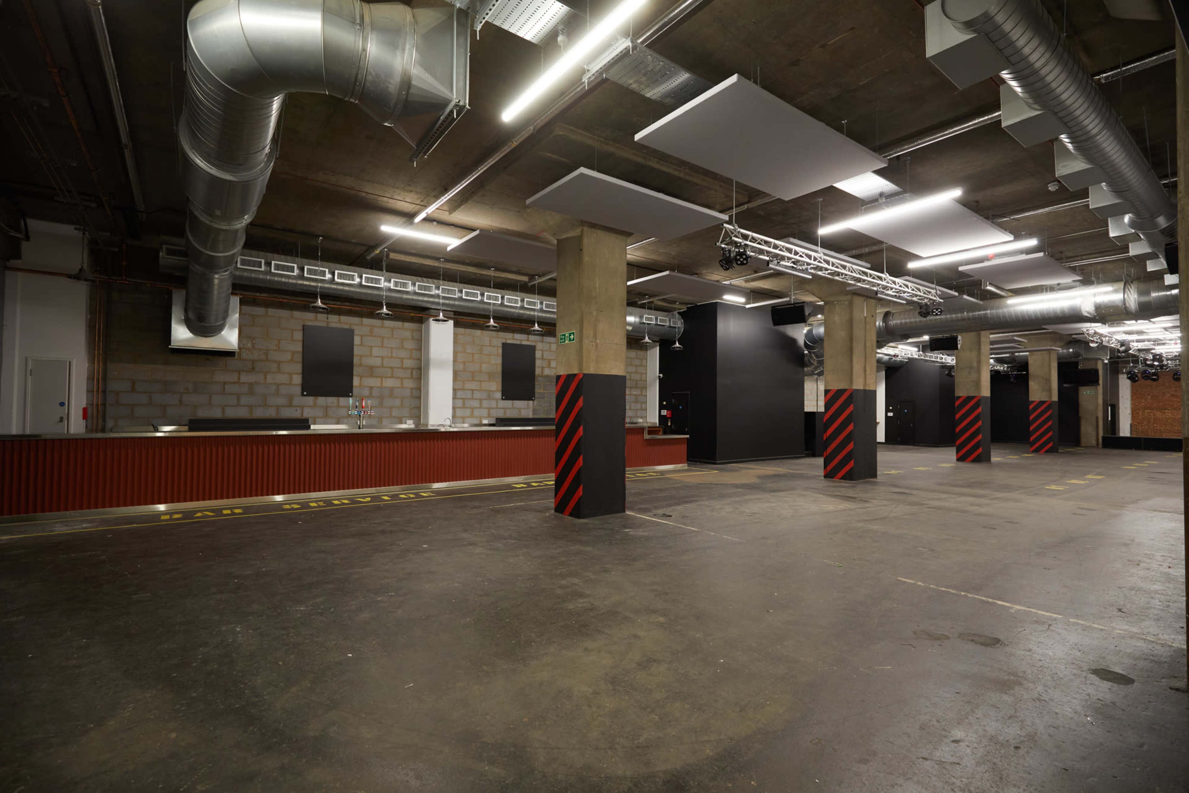 A large, empty industrial space with exposed concrete walls, overhead ventilation ducts, and a bar area with a red and black striped design.