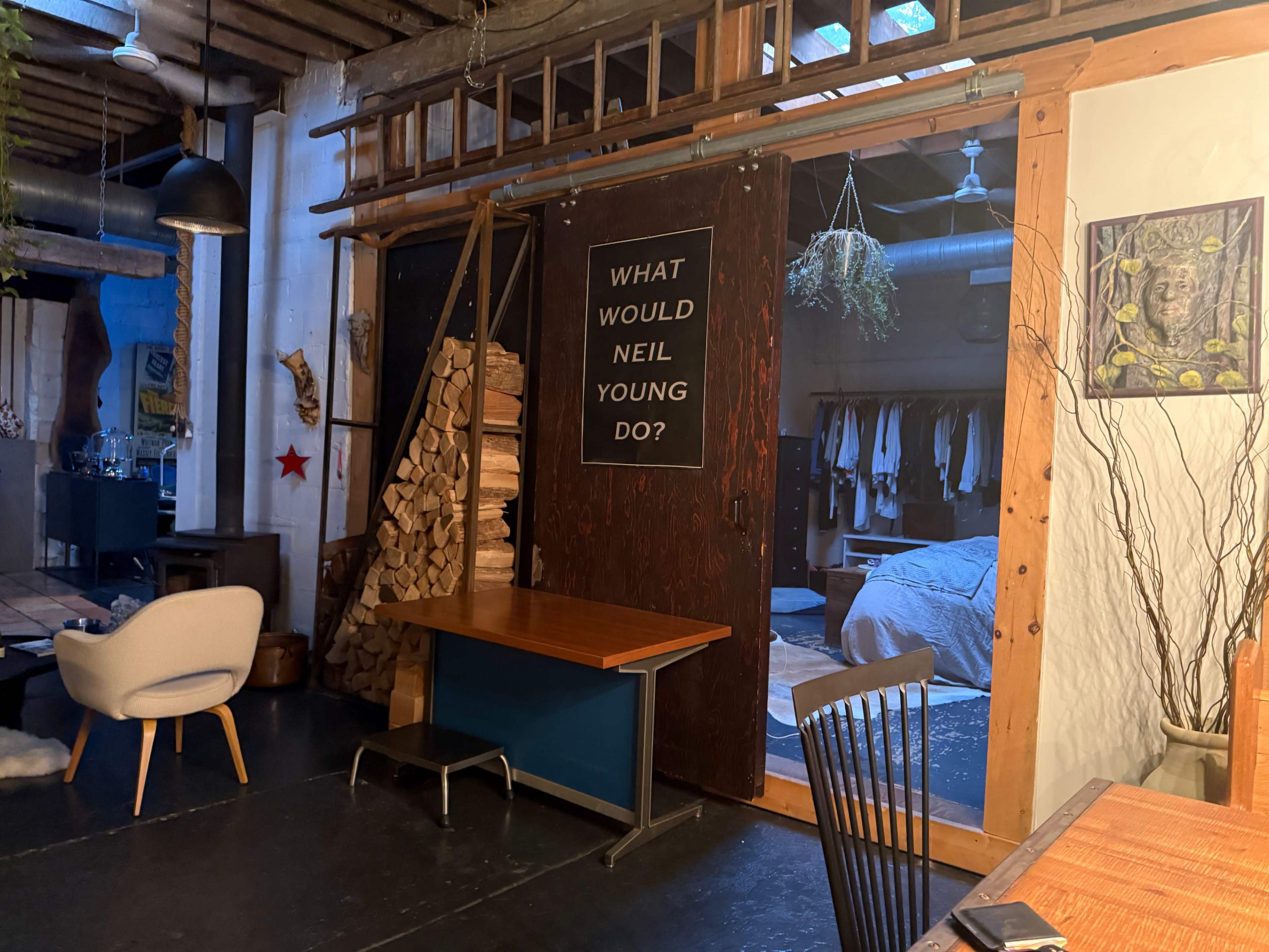 The image shows a cozy, rustic interior with wooden features, a large door displaying the phrase "WHAT WOULD NEIL YOUNG DO?", stacked firewood, and visible clothing hanging in a dimly lit room beyond.