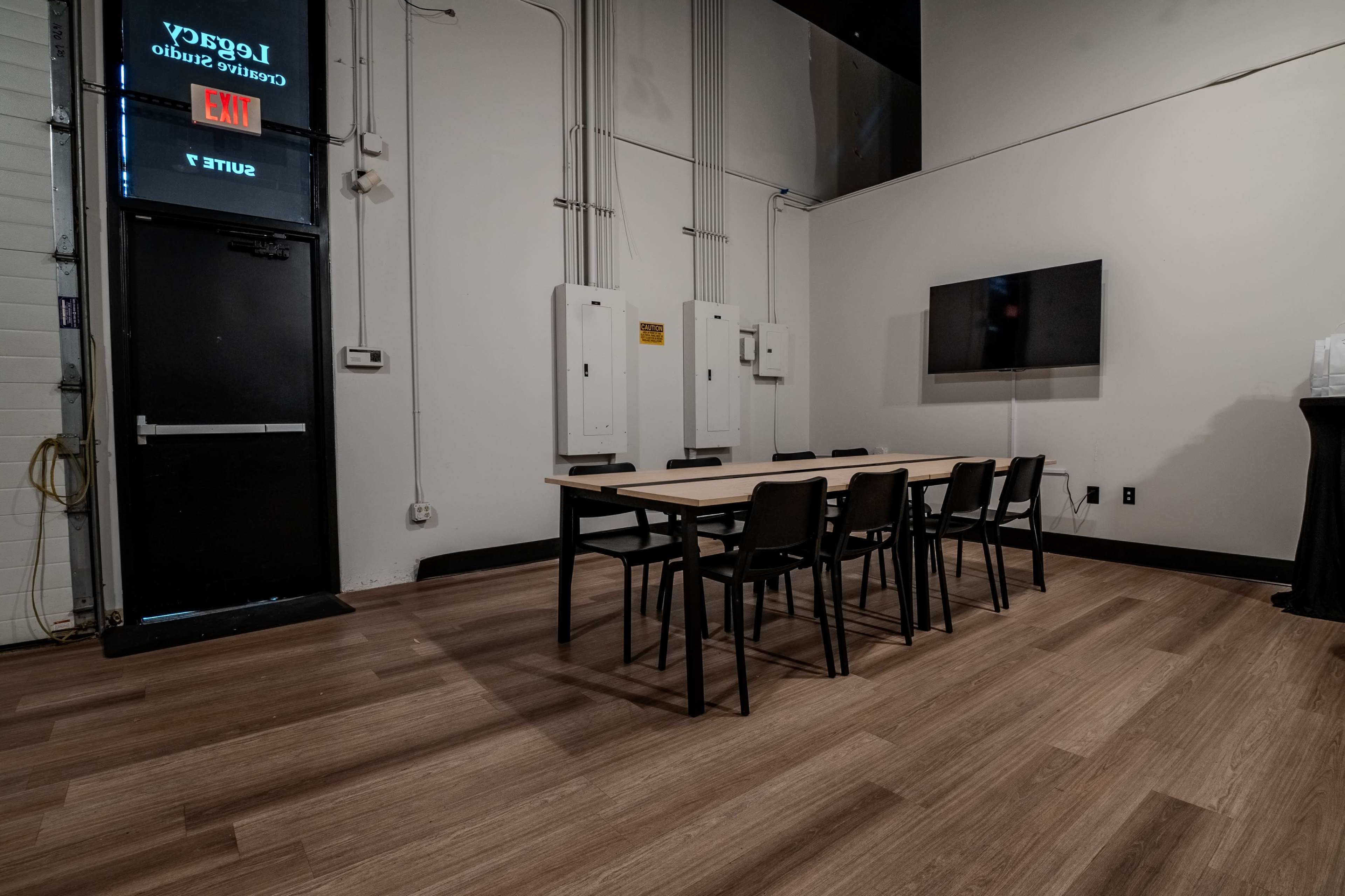 A sparse meeting room features a rectangular table surrounded by six black chairs, with electrical panels and a wall-mounted television in the background.