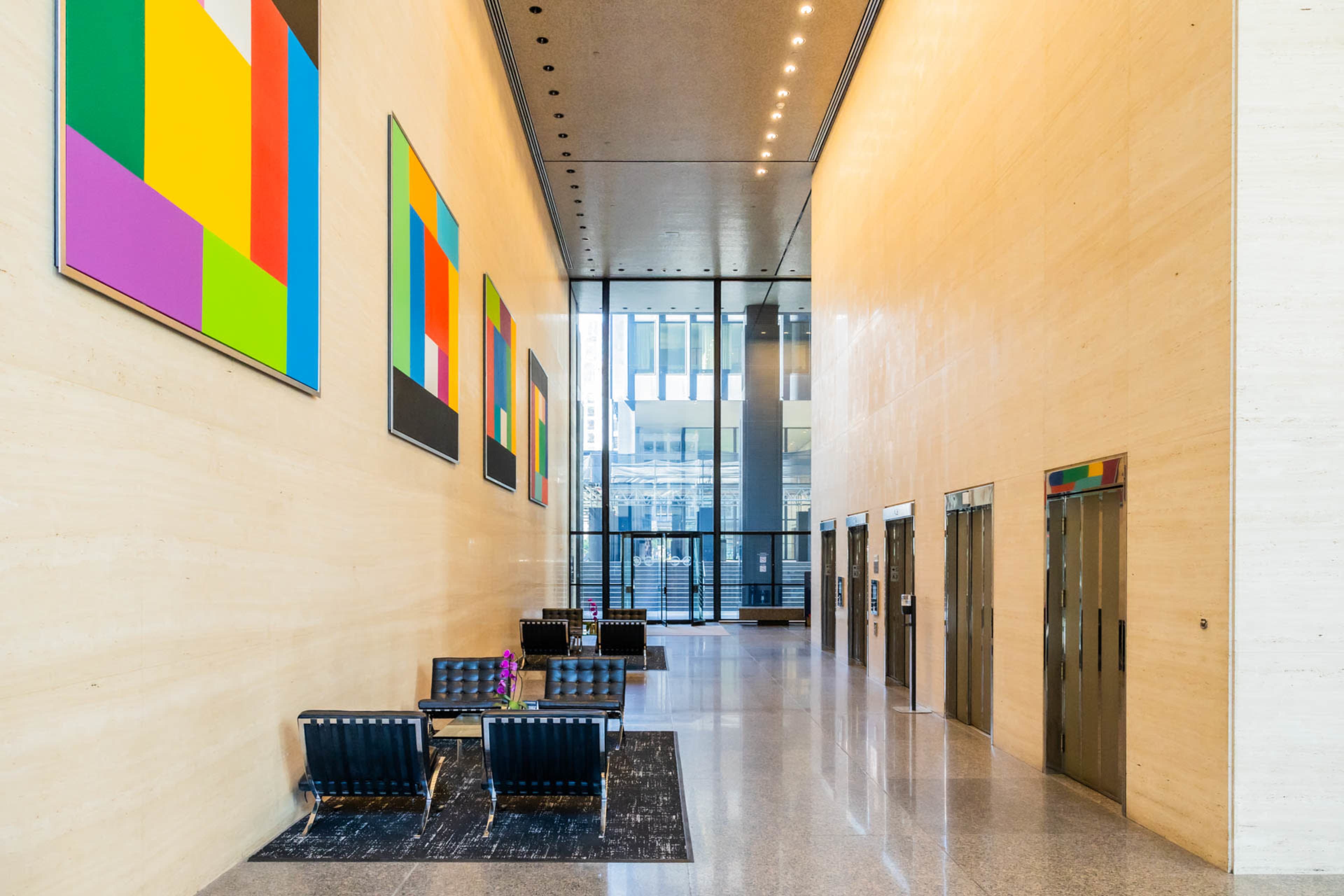 The image shows a modern lobby with a high ceiling, large windows, colorful abstract paintings on the walls, and an arrangement of seating near the elevators.