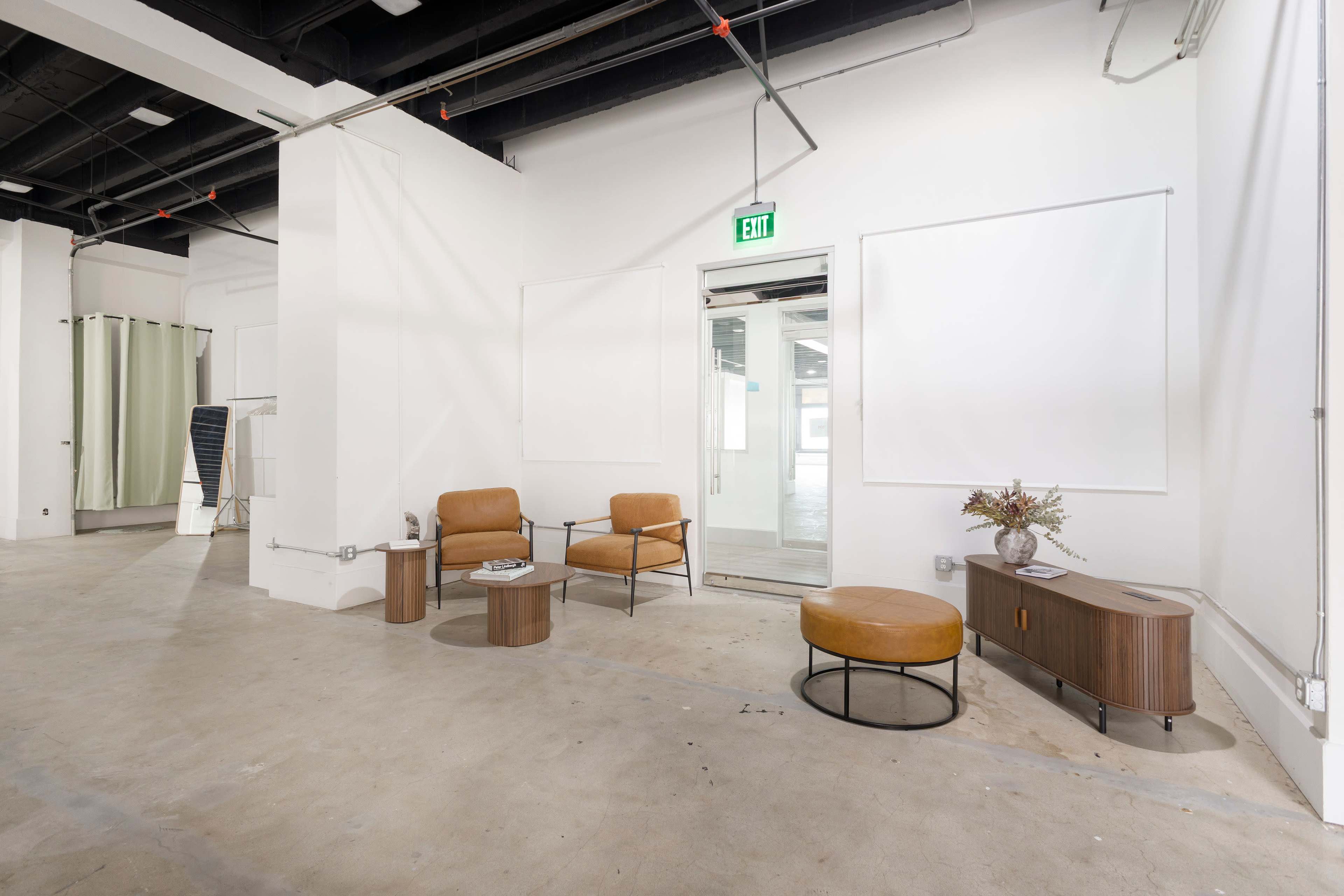 The image shows a minimalist interior space featuring two brown chairs, a round ottoman, and a side table with a decorative vase, all placed against white walls and open to a doorway.