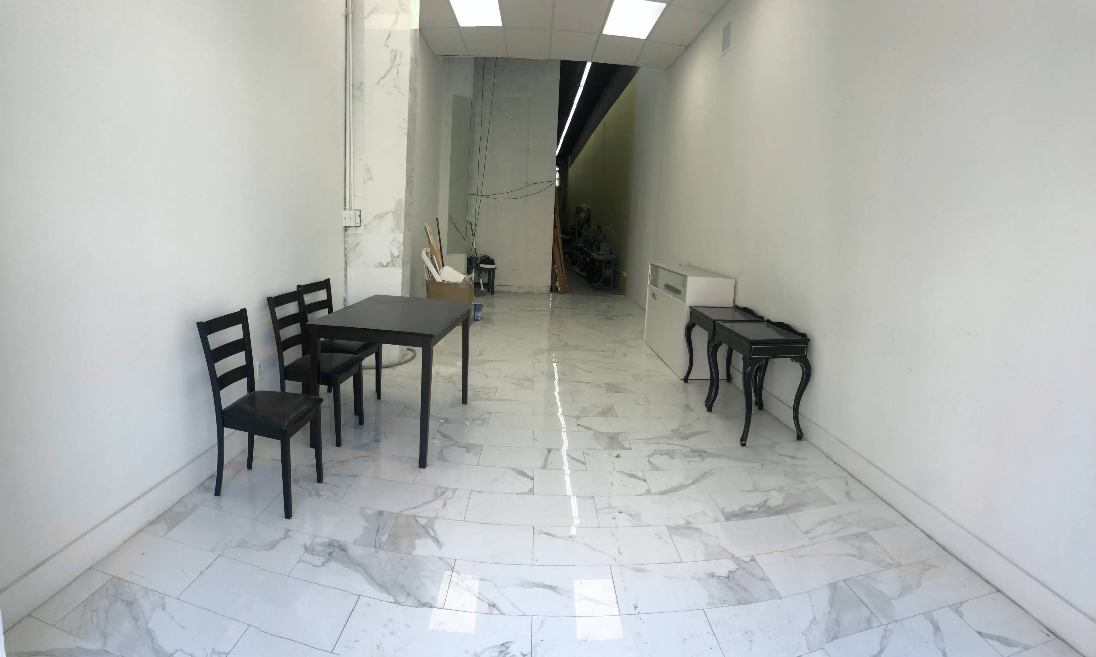 The image shows a spacious, minimally furnished room with a marble floor, featuring a table and chairs on one side and two small tables against the wall.