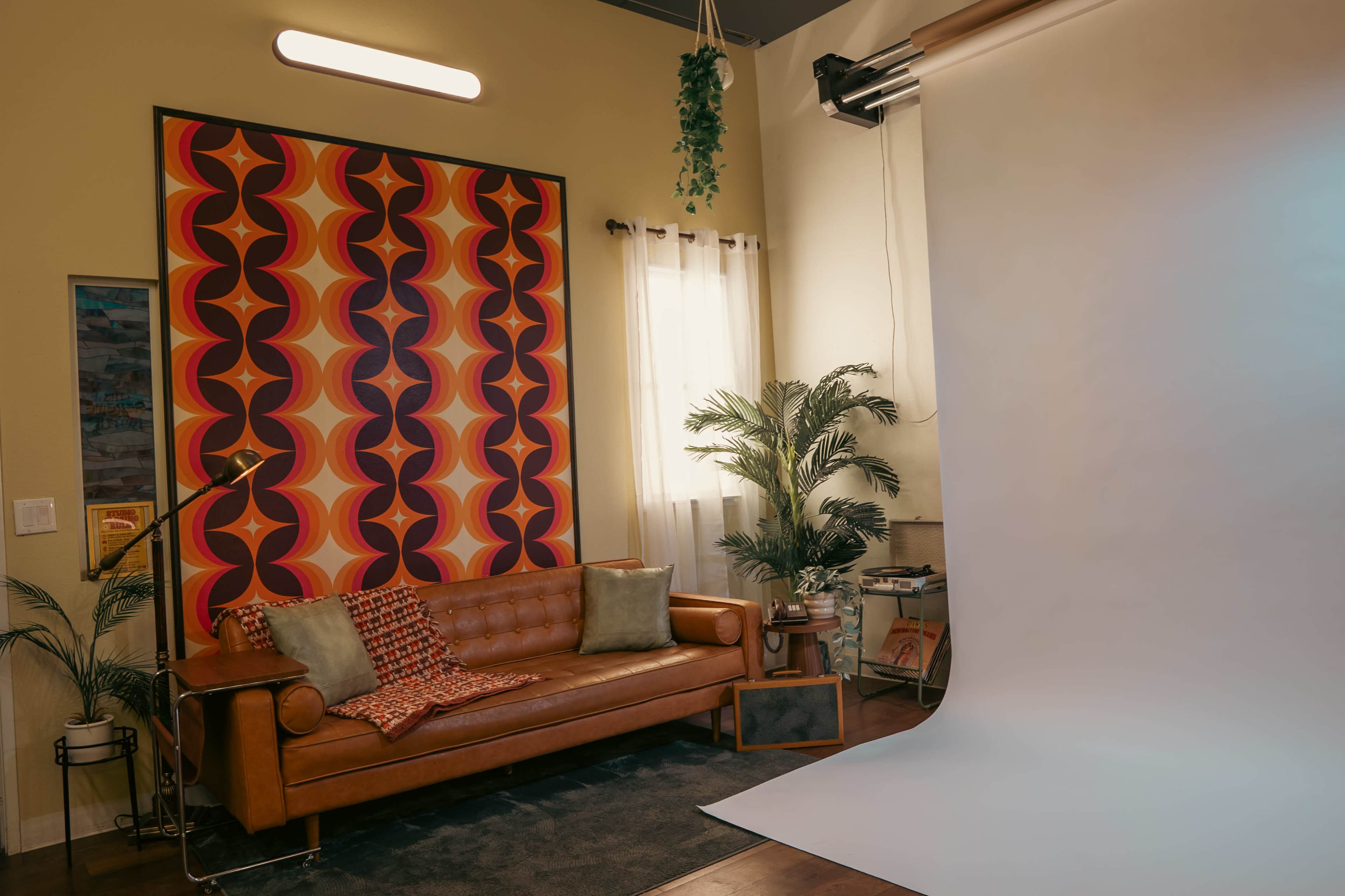 The image shows a stylish living space featuring a brown leather sofa, vibrant patterned wall art, and a plain backdrop next to a window with natural light.
