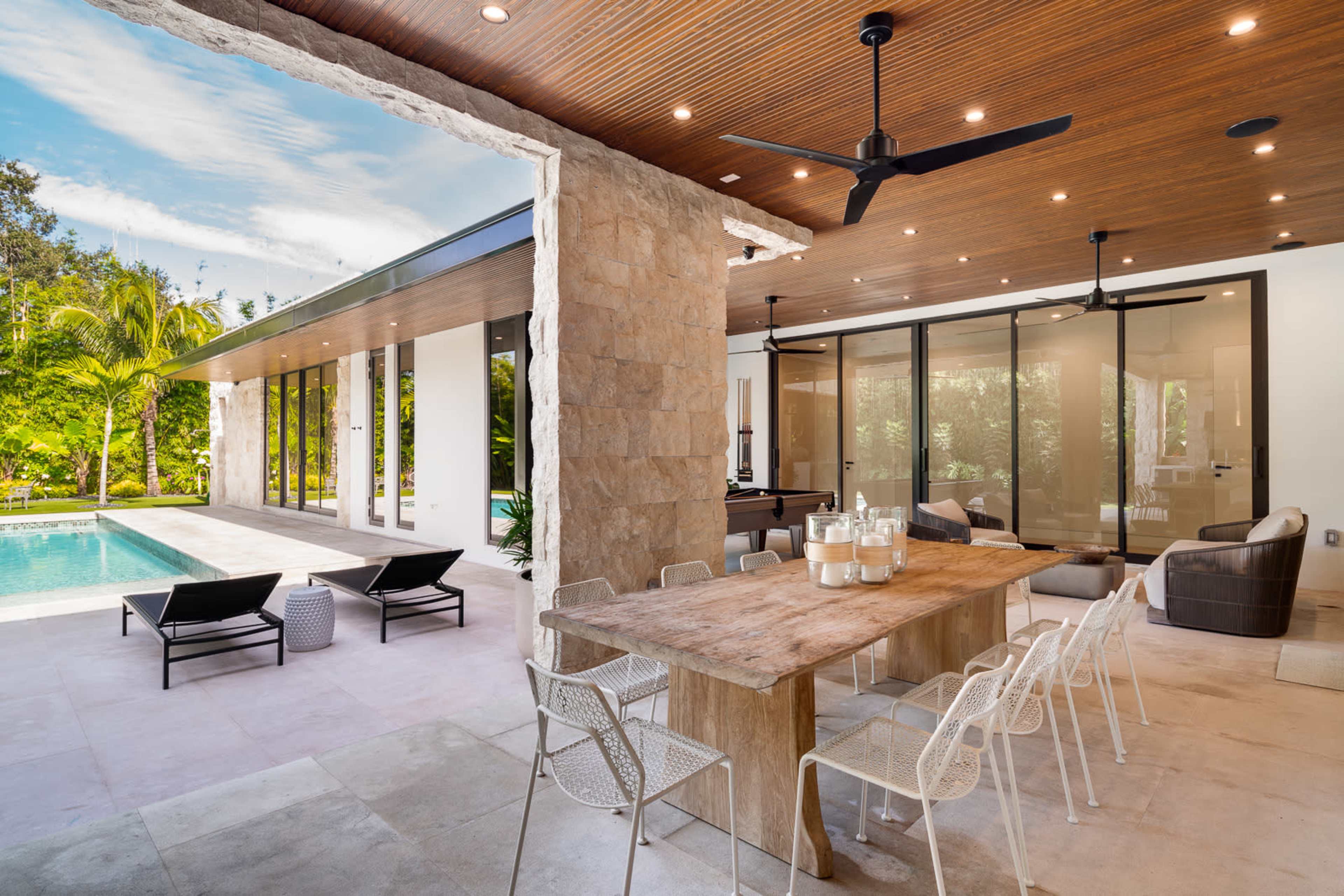 The image shows a modern outdoor dining area adjacent to a swimming pool, featuring a wooden table and white metal chairs under a canopy with ceiling fans.