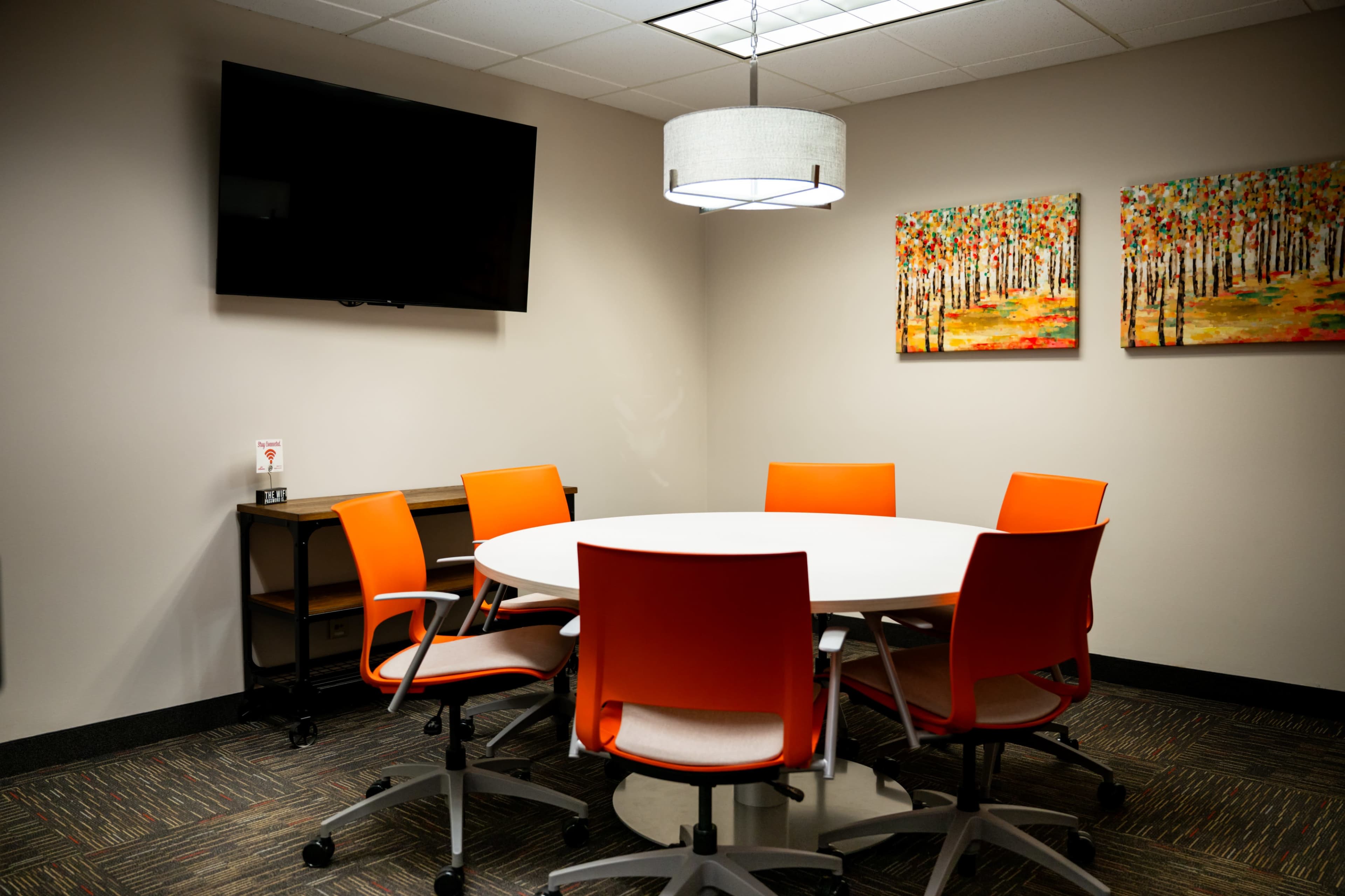 The image shows a modern conference room featuring a round white table surrounded by six orange chairs, a flat-screen TV on the wall, and two colorful paintings of trees.