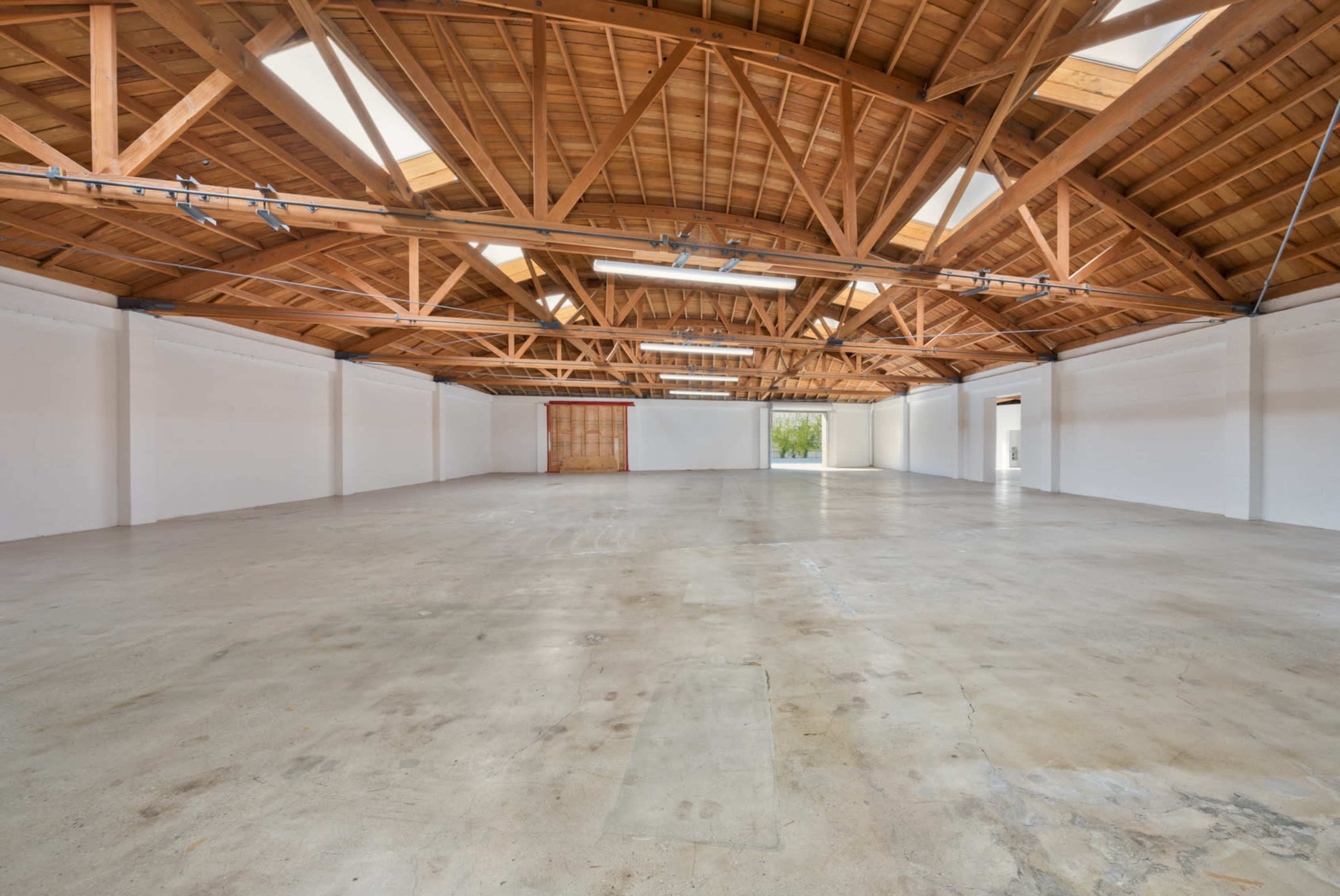 The image shows a spacious, empty warehouse with a high wooden ceiling and large windows along one side.