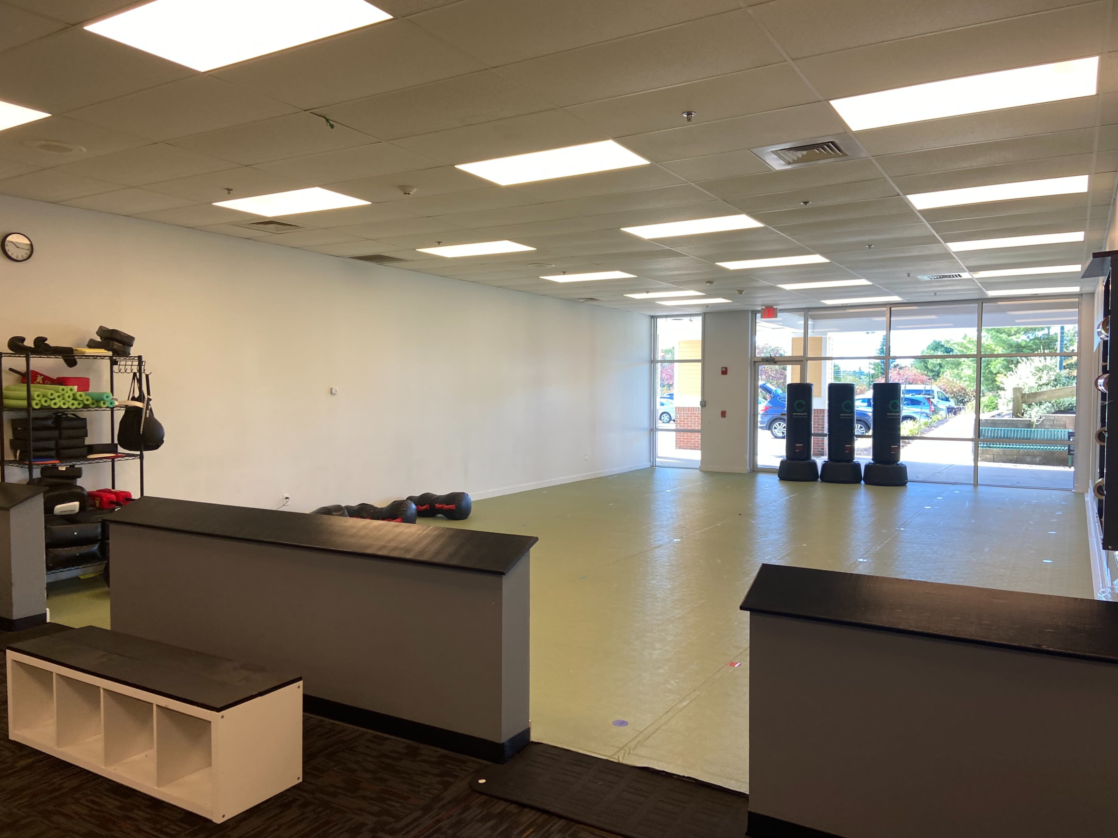 A spacious, empty room with a light-colored floor, large windows, and fitness equipment along one wall.