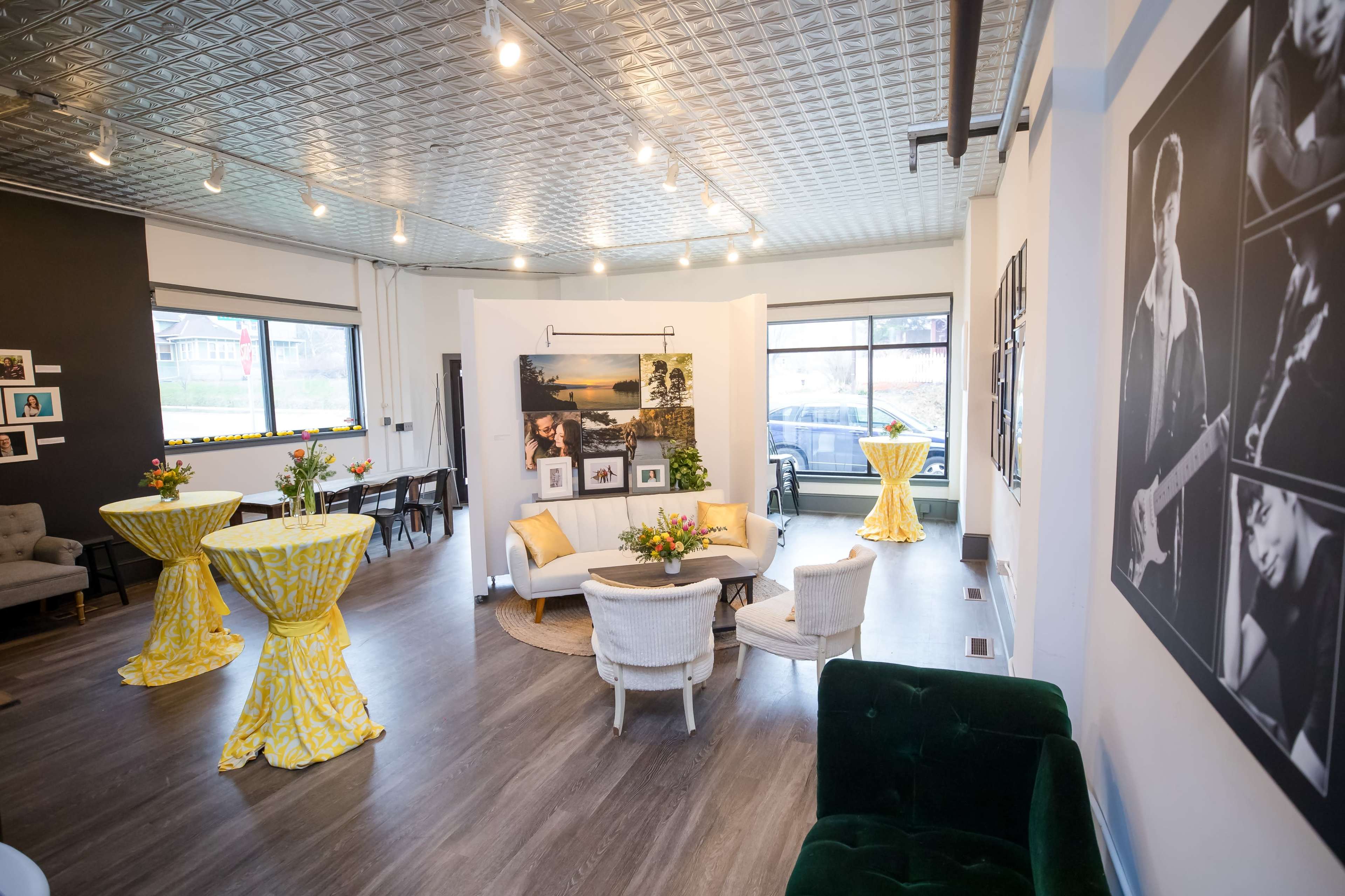 A modern event space with a seating area featuring a white sofa and coffee table, surrounded by tall tables covered with yellow tablecloths, and framed black-and-white photographs on the walls.