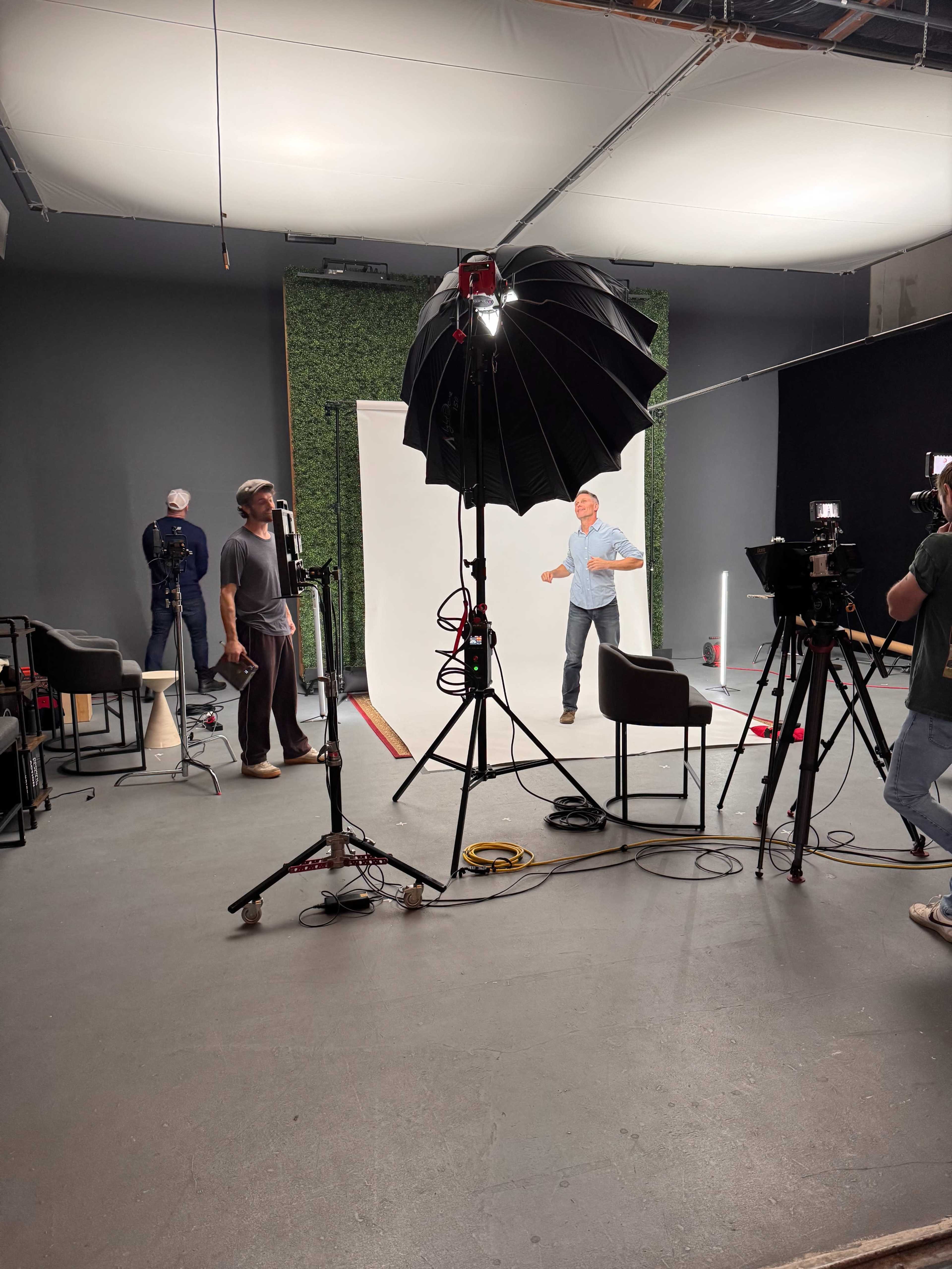 A film studio setup features a man standing in front of a white backdrop, with lighting equipment, cameras, and crew members preparing for a shoot.