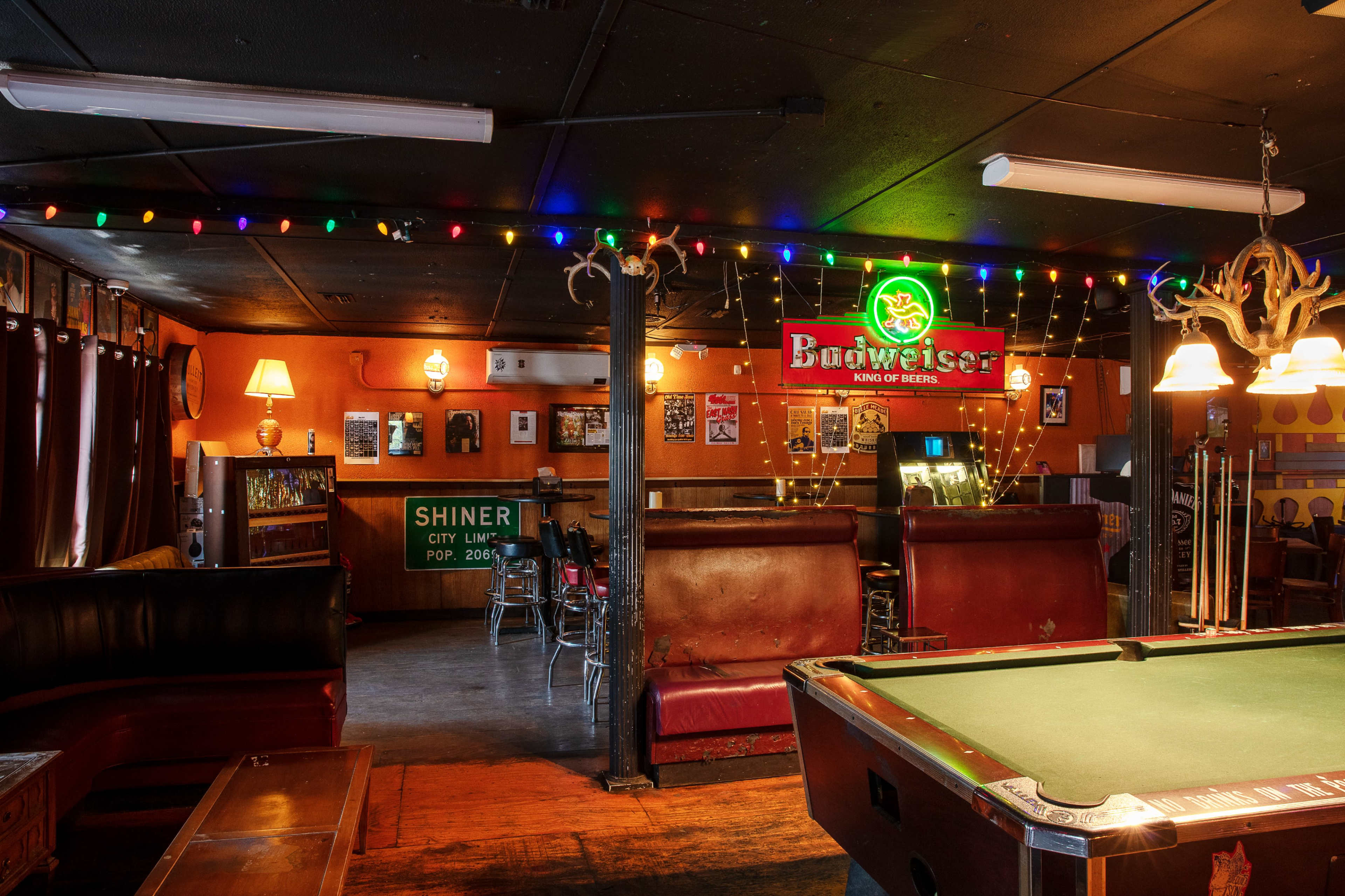 The image shows a dimly lit bar interior with a pool table, colorful string lights, and a Budweiser neon sign.