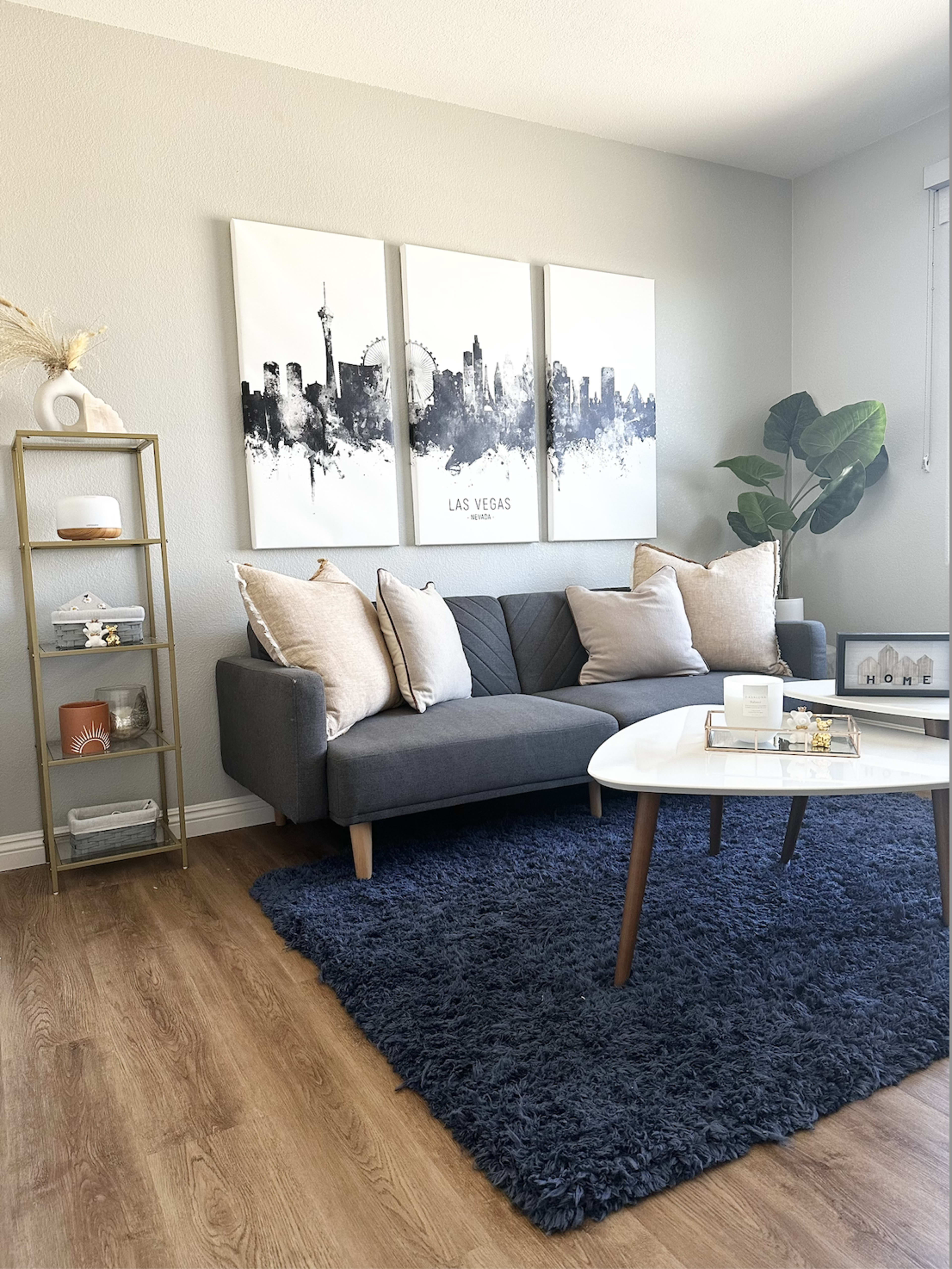 The image shows a modern living room featuring a gray sofa with decorative pillows, a coffee table, a shelf with decor items, a large wall art piece of a city skyline, and a potted plant.