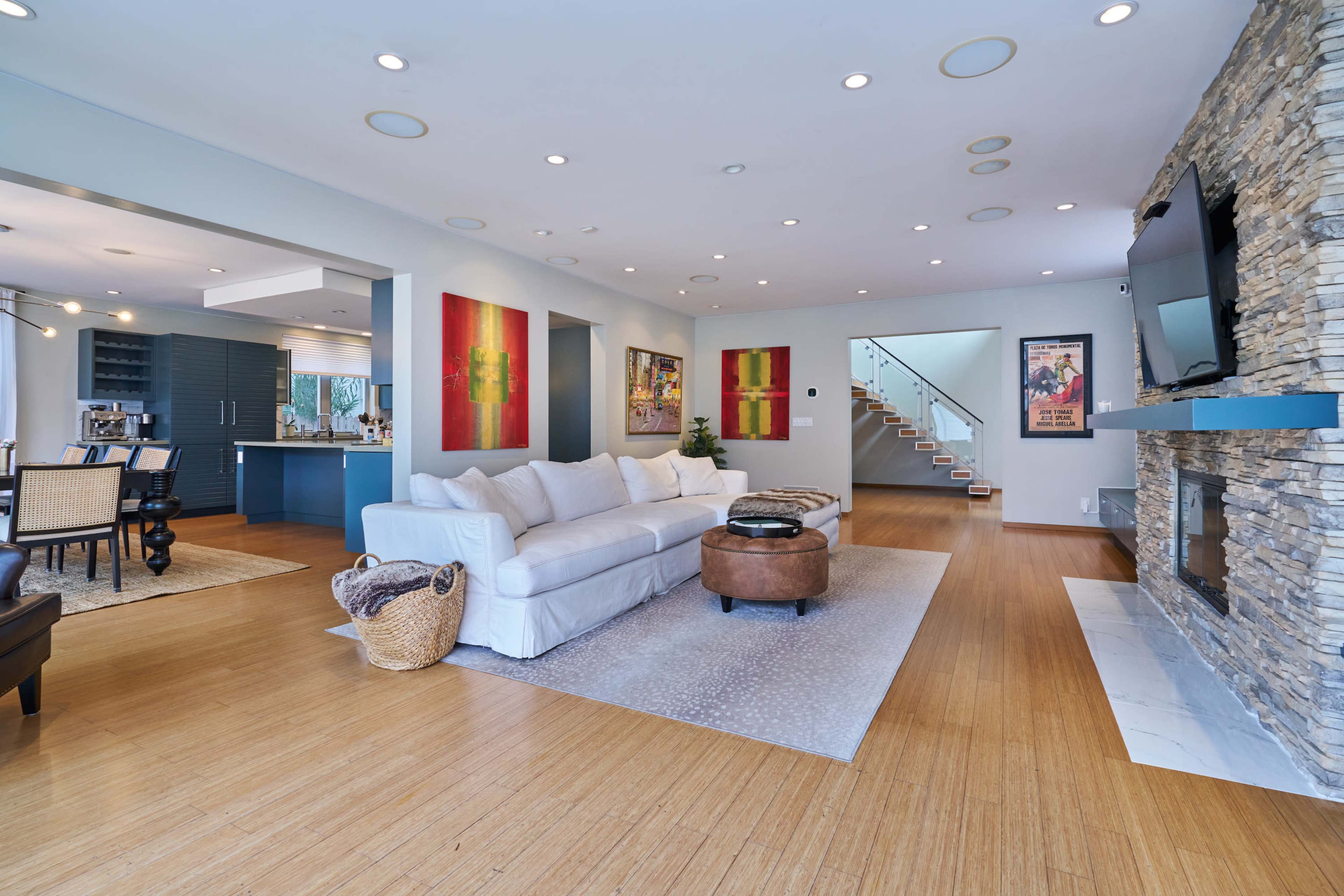 The image shows a modern living room with a white couch, stone fireplace, and large windows, featuring artwork on the walls and a staircase in the background.