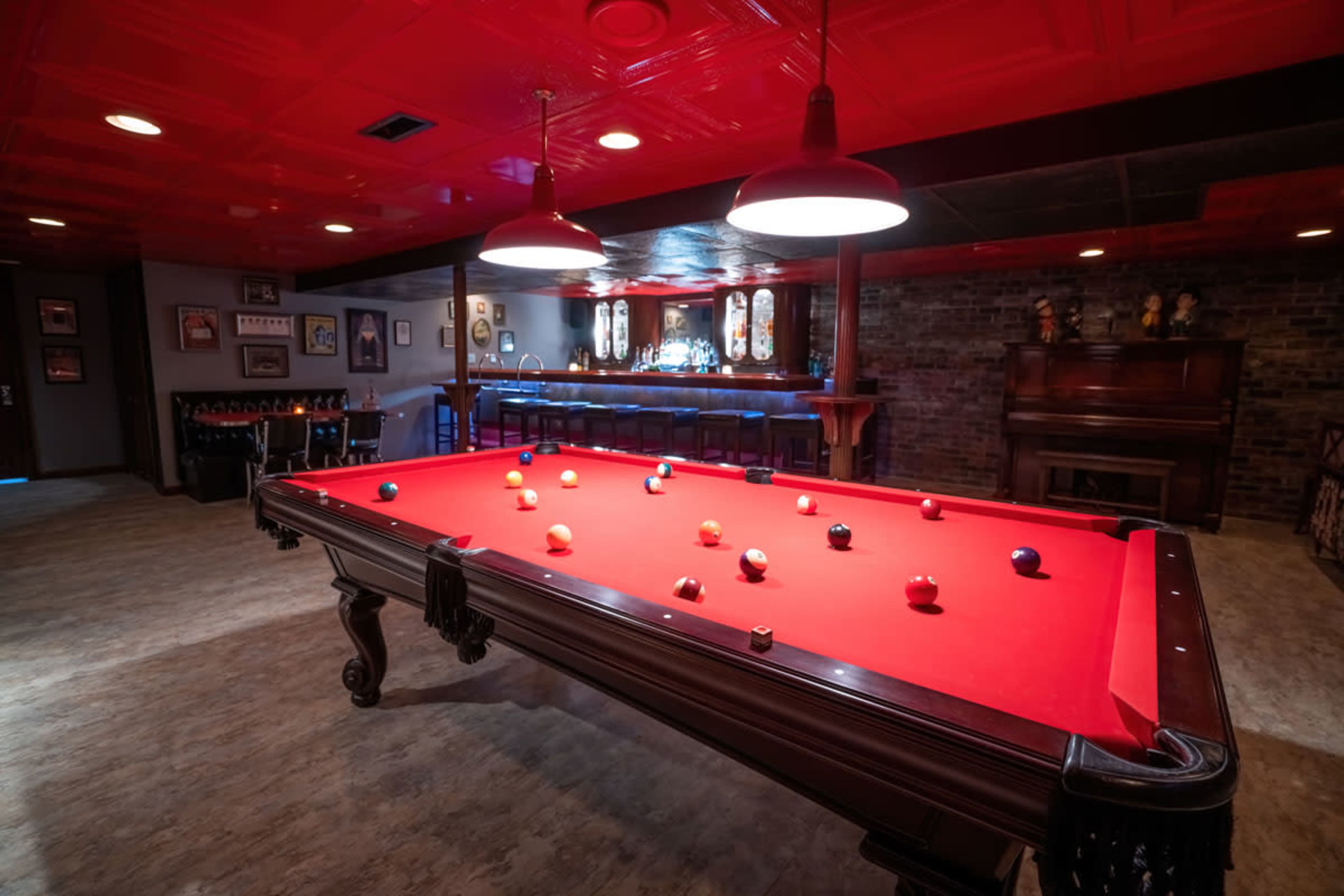 A billiards room featuring a red felt pool table with balls arranged on it, surrounded by dim lighting and a bar area in the background.