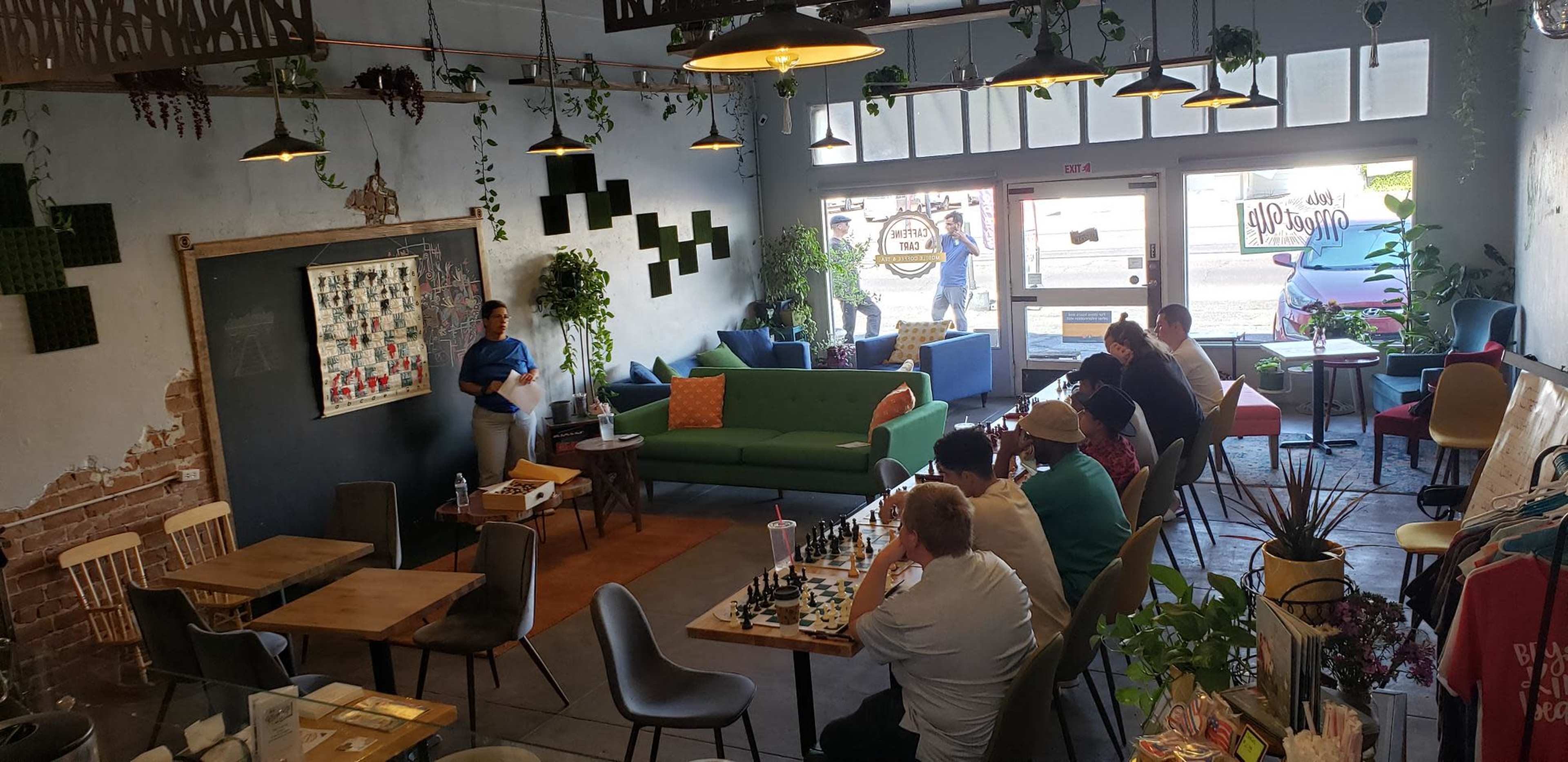 A cozy café features a chess area with several players, seating arrangements, and plants, and a person is standing by a chalkboard at the front.