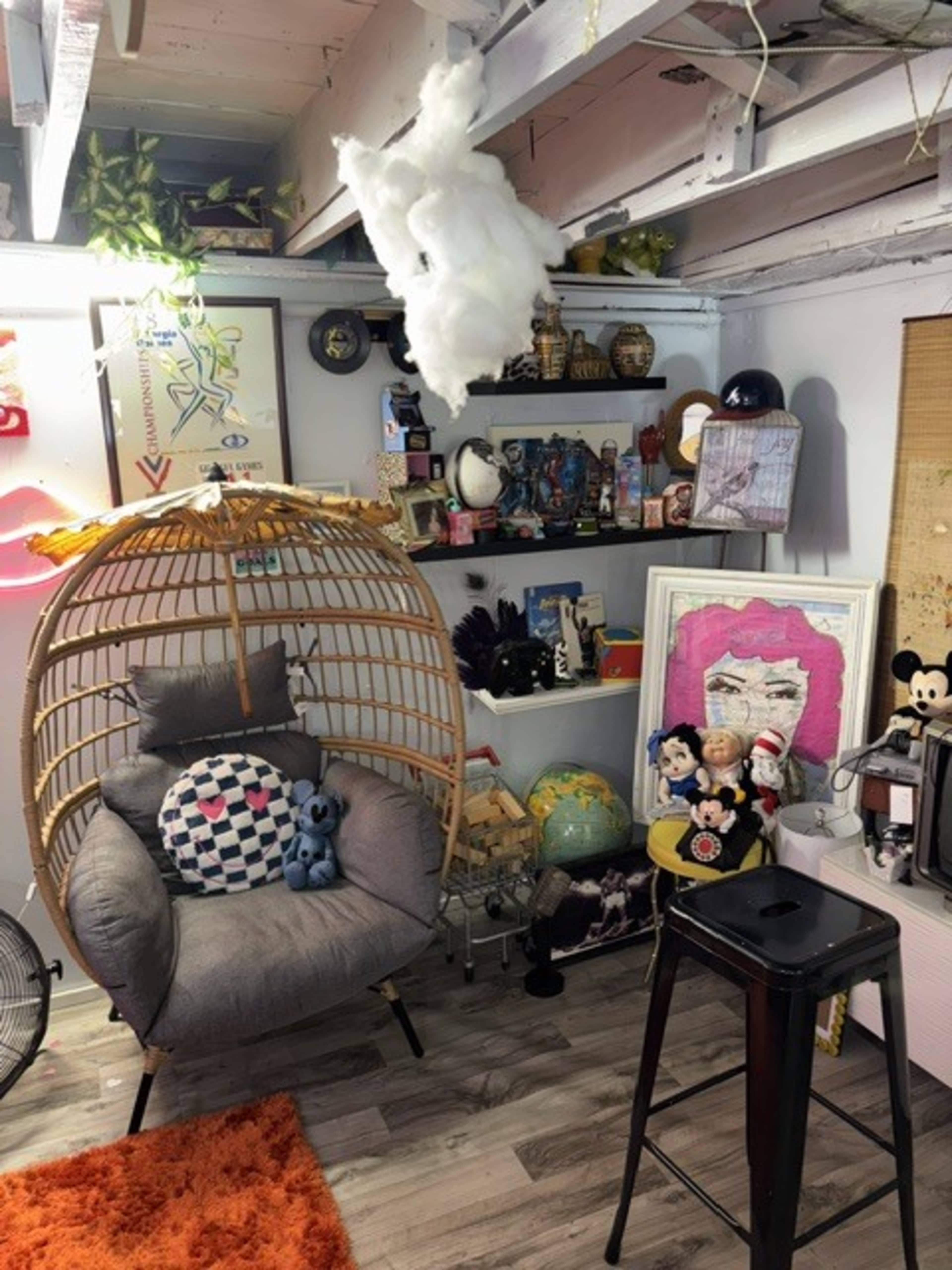 The scene features a cozy corner with a rattan hanging chair, shelves filled with various decorative items, and a small orange rug on the floor.