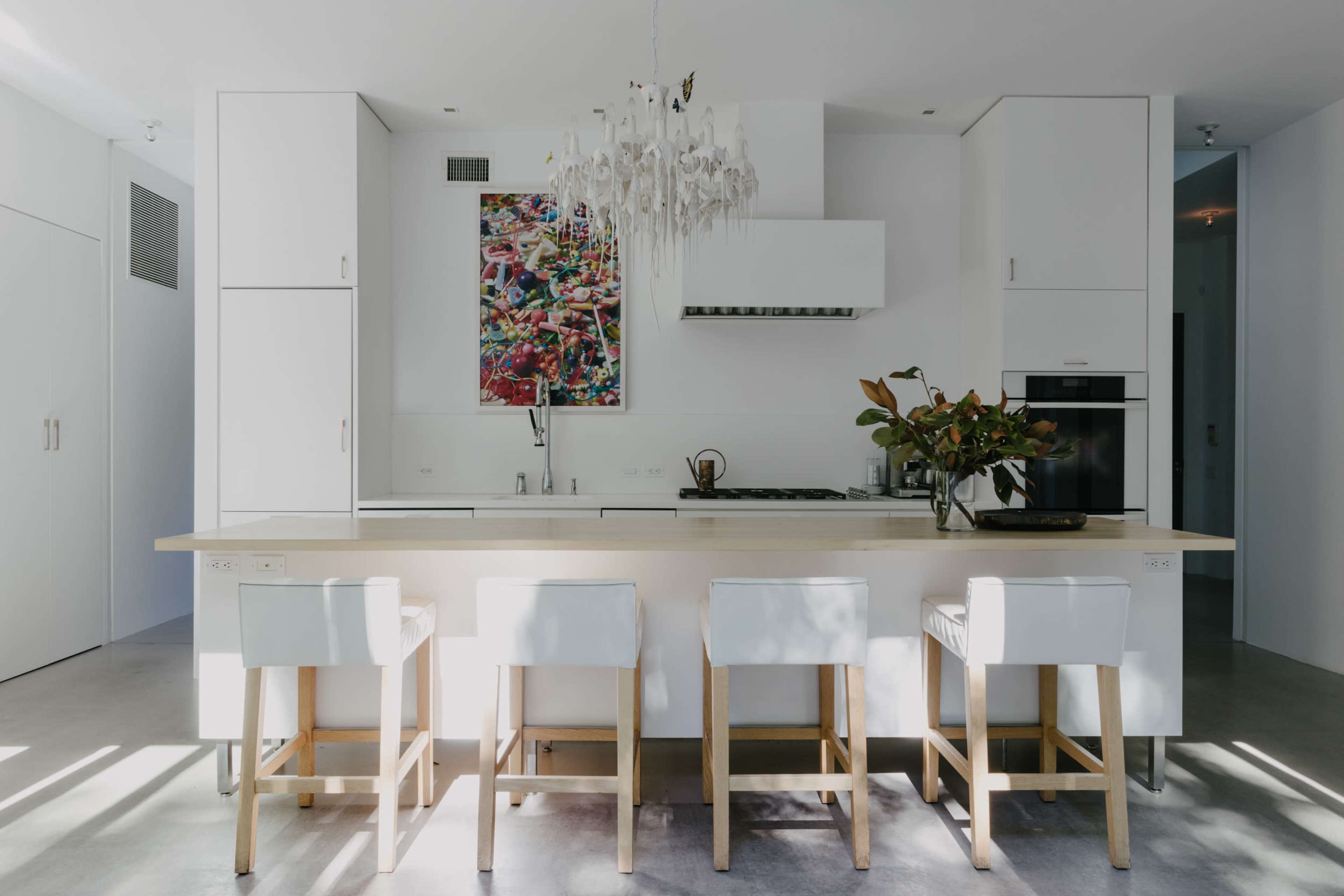 A modern kitchen features a central island with four white stools, a colorful artwork on the wall, and sleek cabinetry.