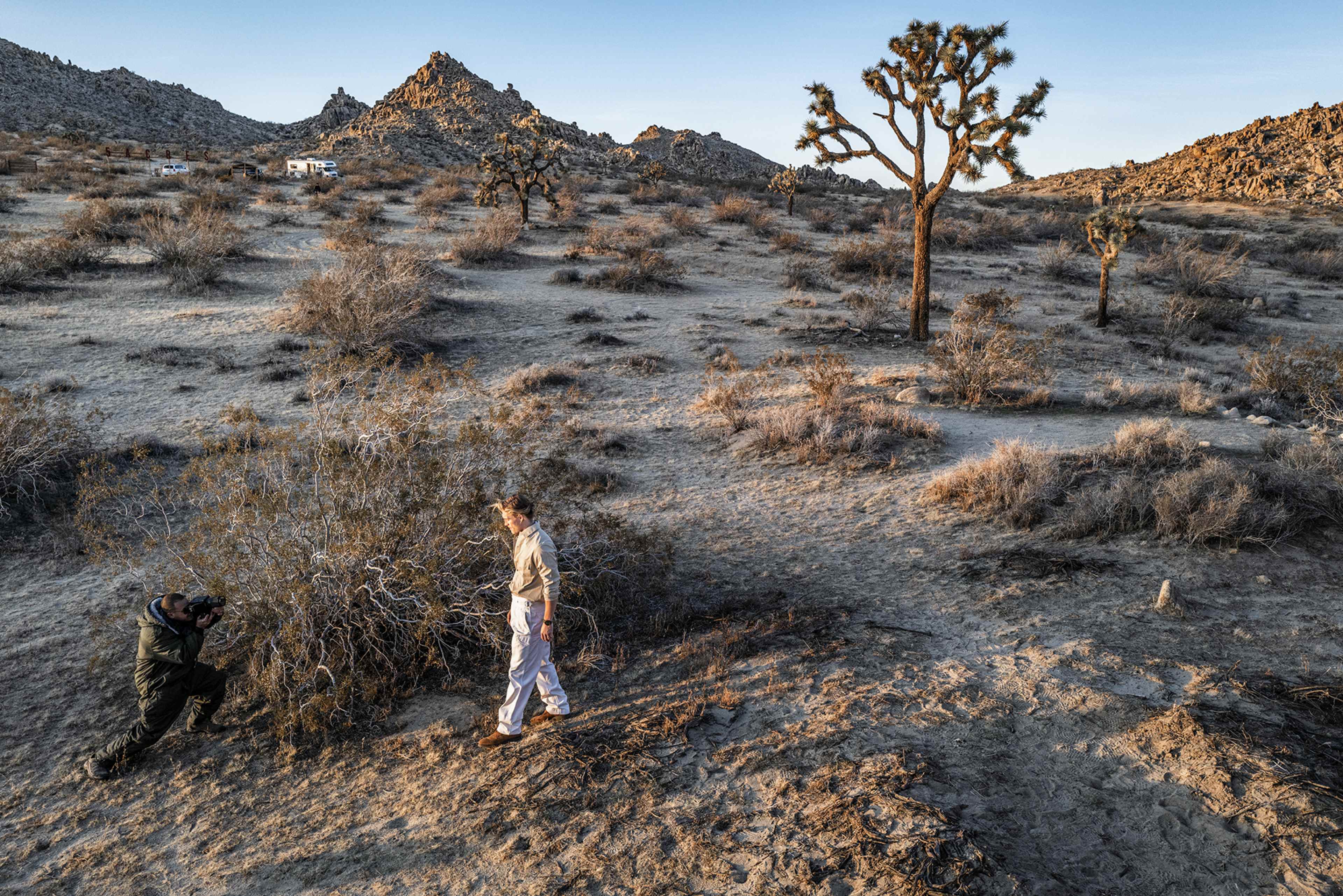 A person poses for a photograph in a desert landscape with scattered shrubs and rocky mountains in the background.