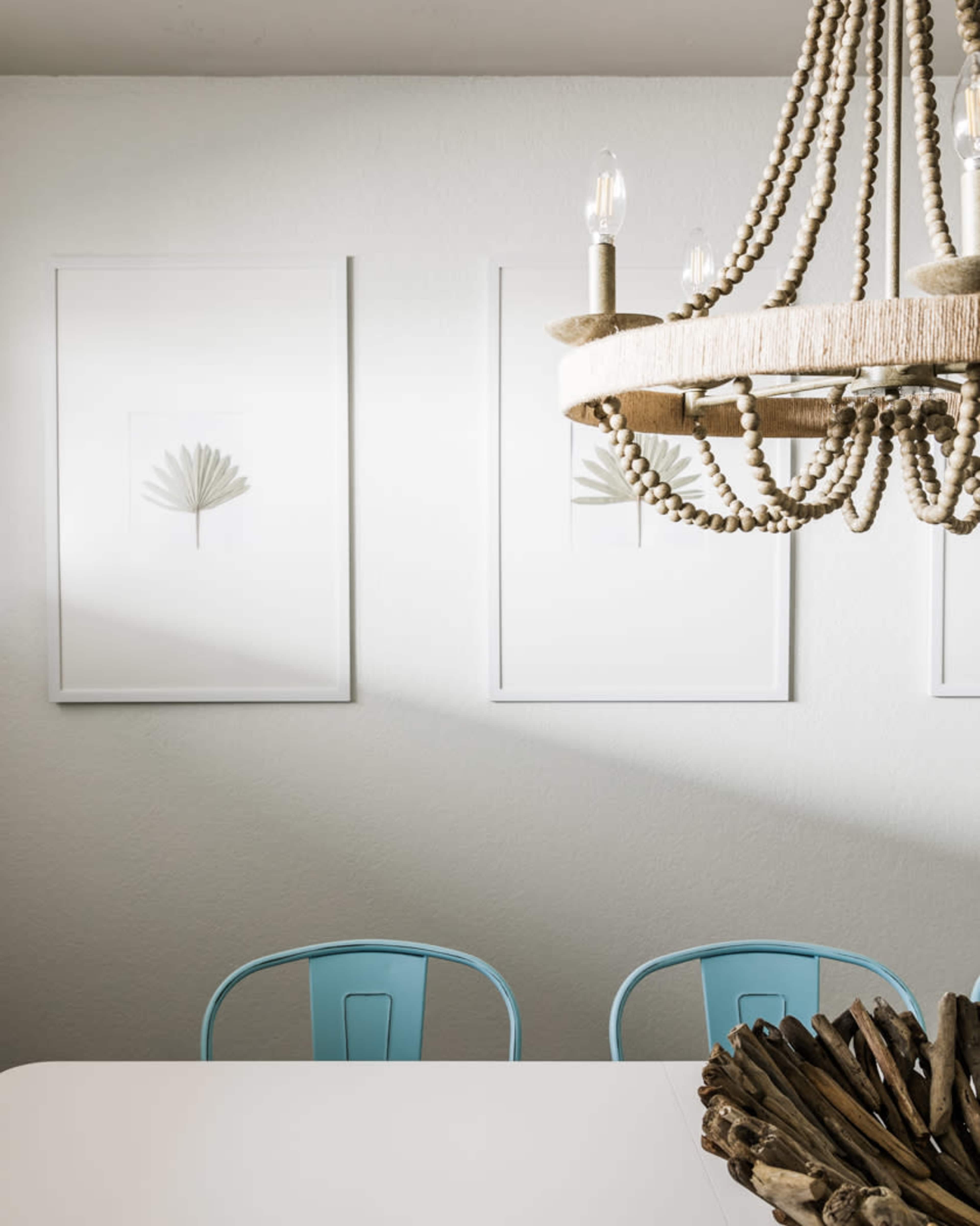 A dining area features a white table with blue chairs, framed artwork on the walls, and a chandelier made of beads and wood.