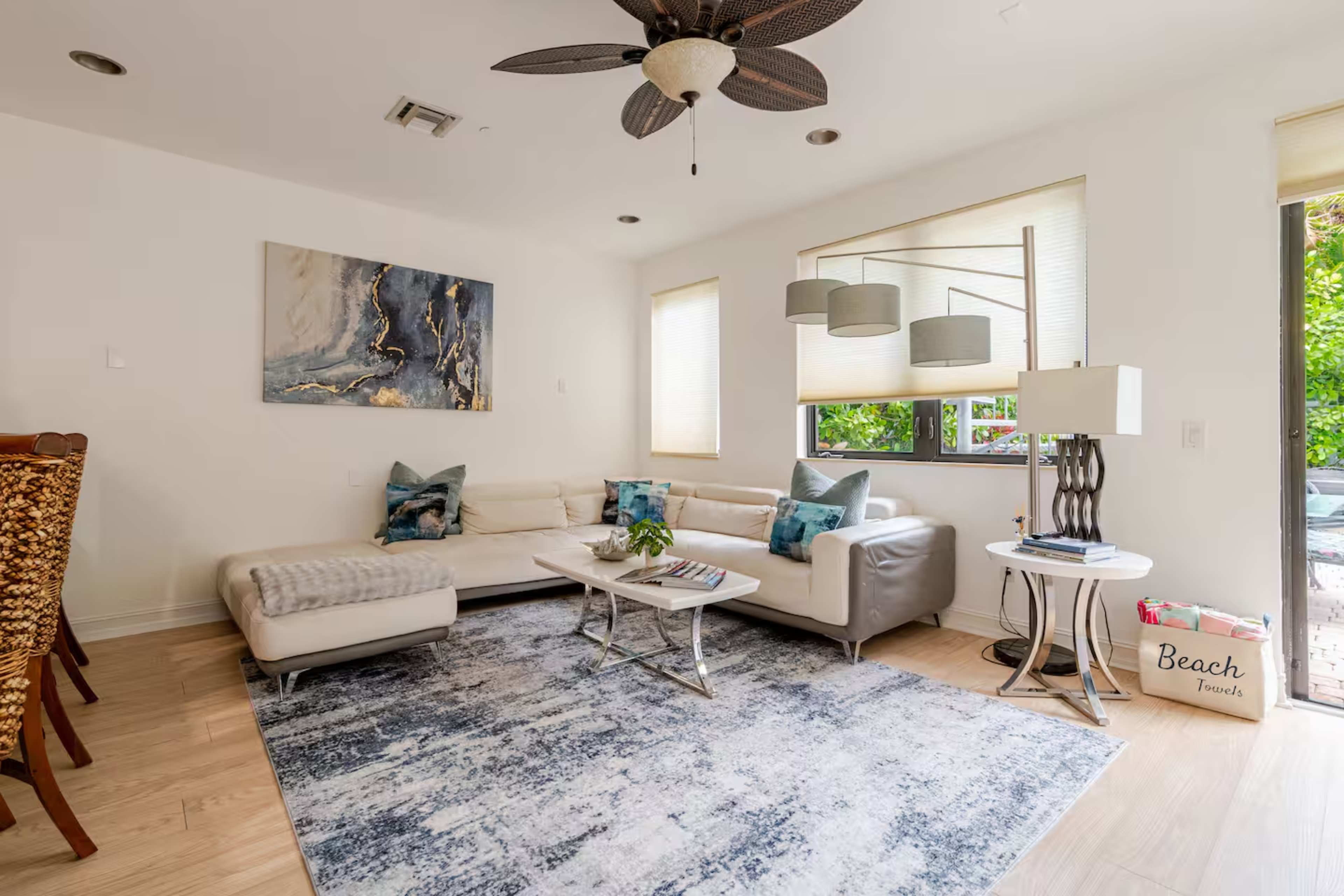 The image shows a modern living room featuring a sectional sofa, a coffee table, and large windows with natural light, along with a dining area visible in the background.