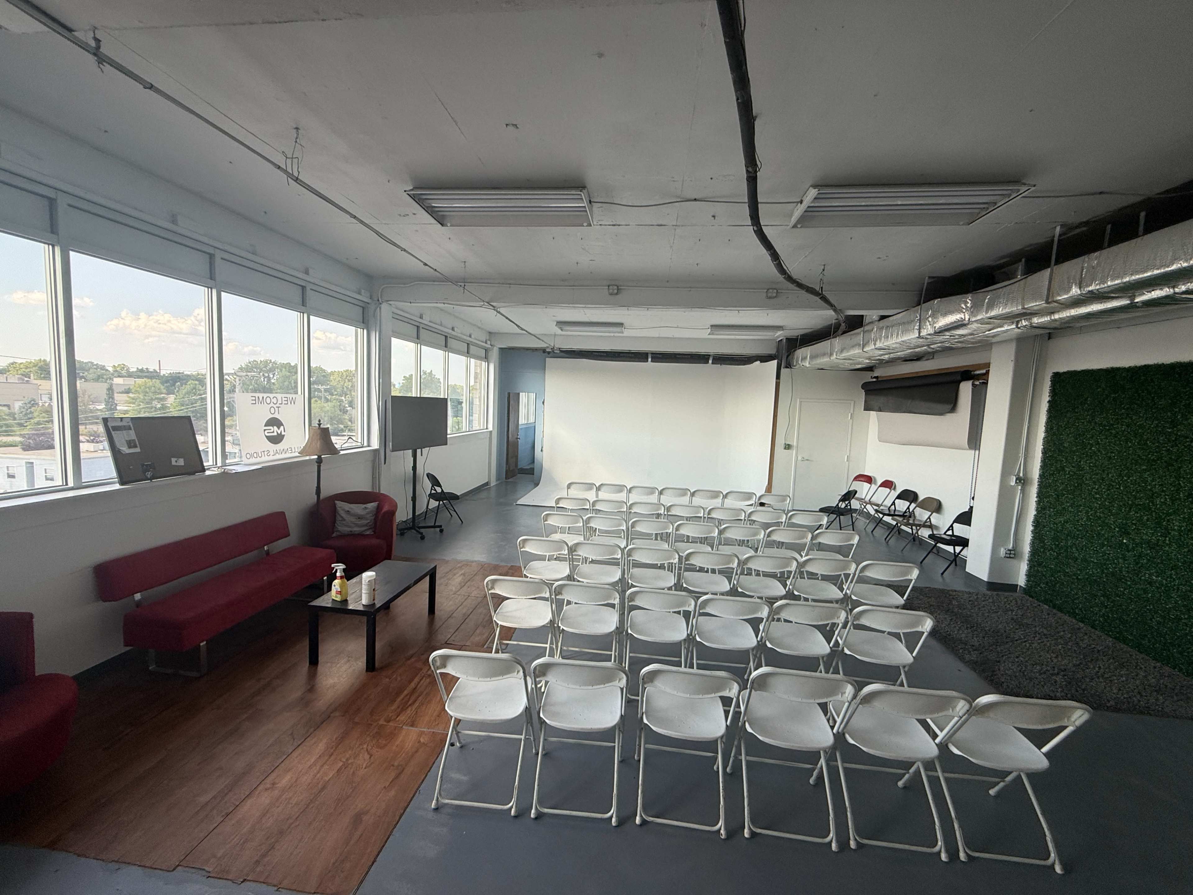 The image shows a spacious, bright room set up with rows of folding chairs facing a blank wall, accompanied by a red sofa and a TV screen on a stand.