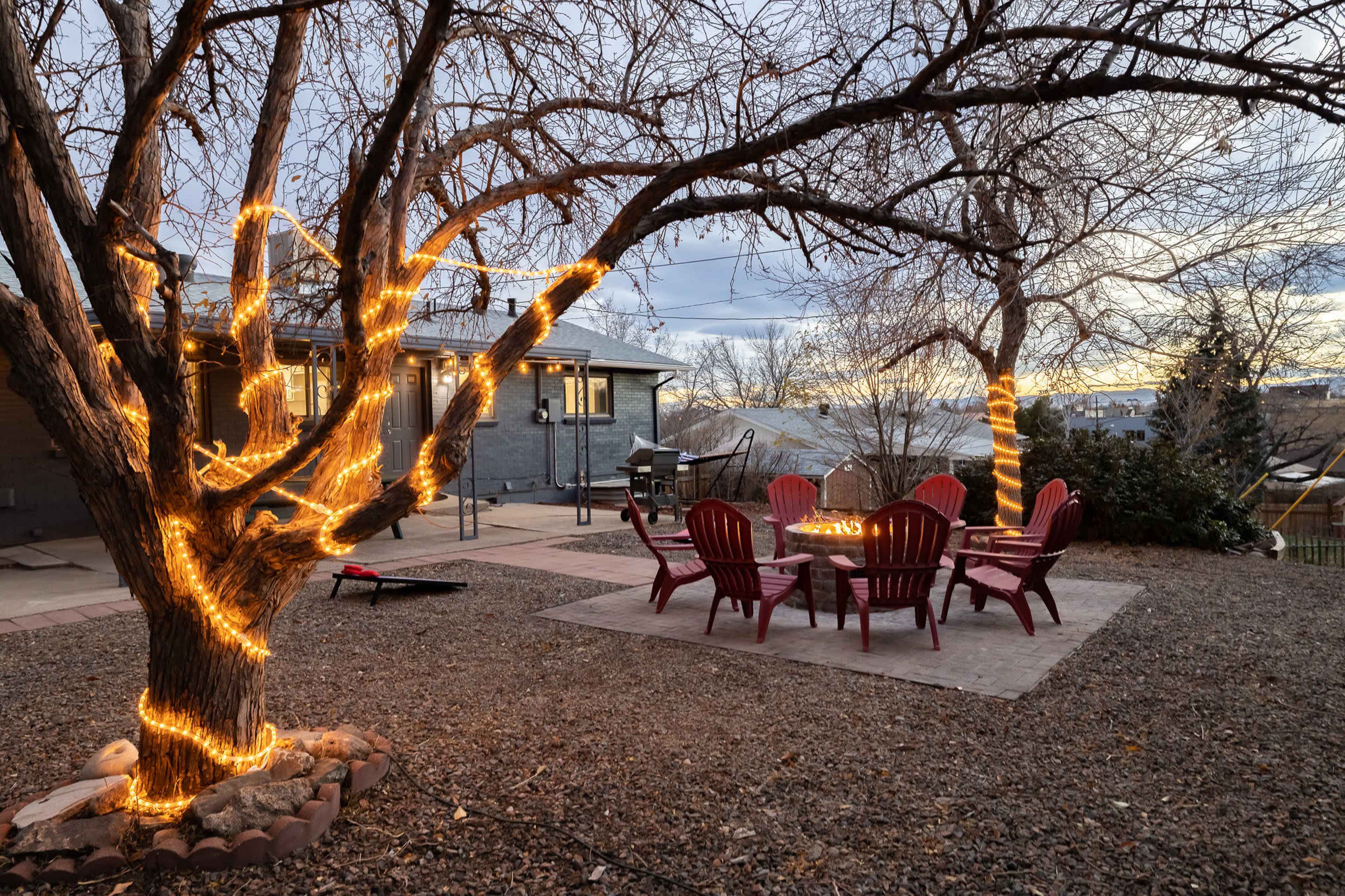 A tree wrapped in lights stands beside a stone patio featuring a circular arrangement of red Adirondack chairs around a fire pit.