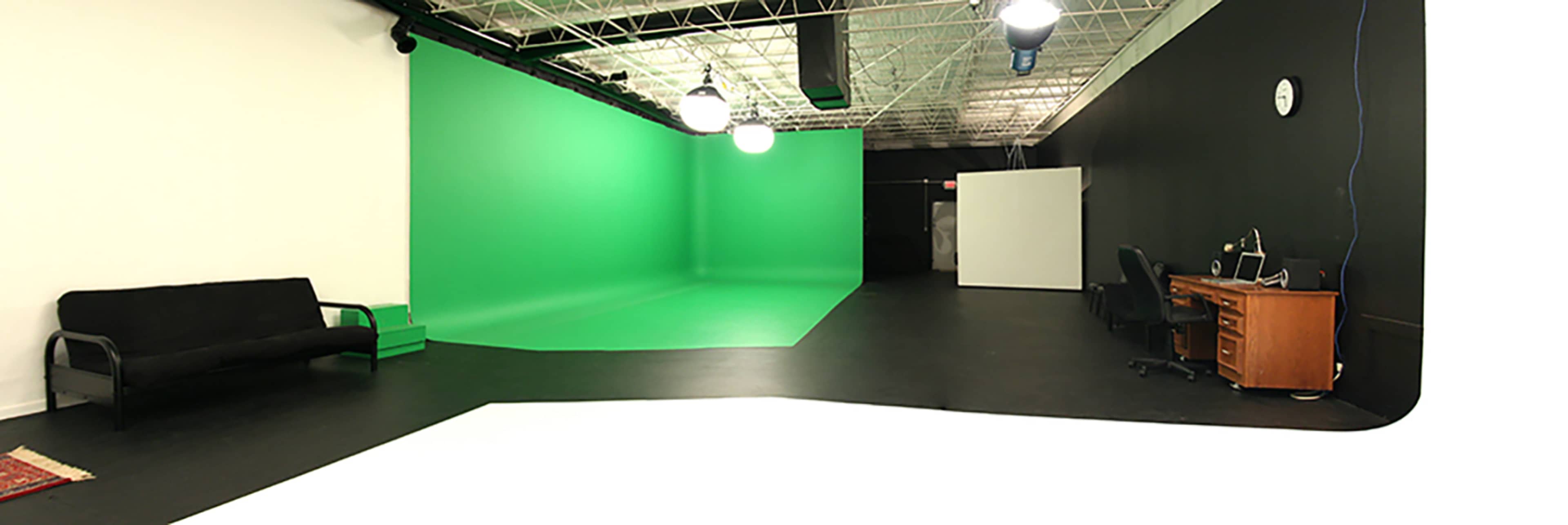 The image shows a studio space featuring a green screen backdrop, a black wall, a desk with a chair, and a black couch.