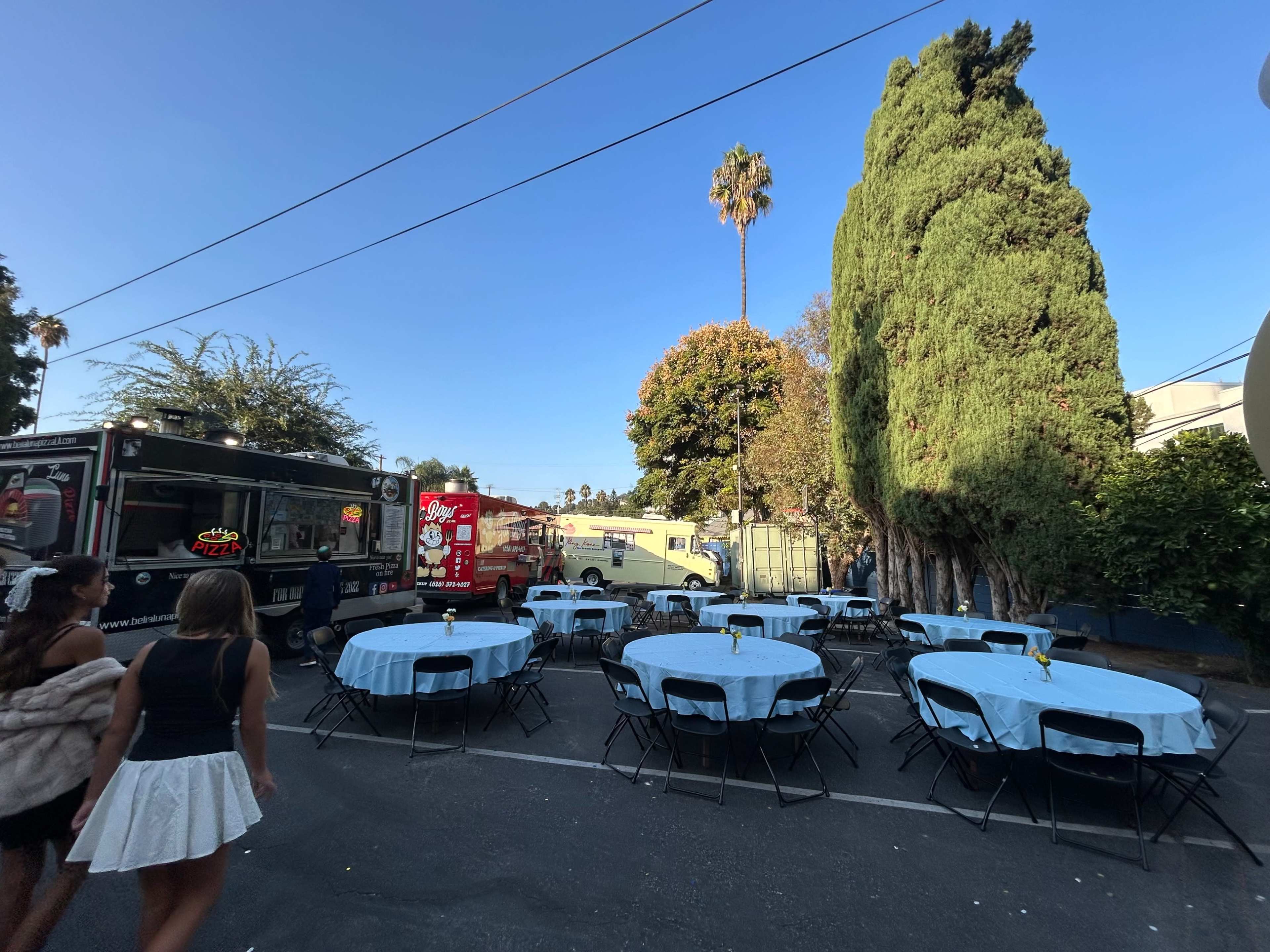 The scene features an outdoor event space with tables covered in light blue tablecloths and food trucks parked nearby, surrounded by palm trees and greenery.