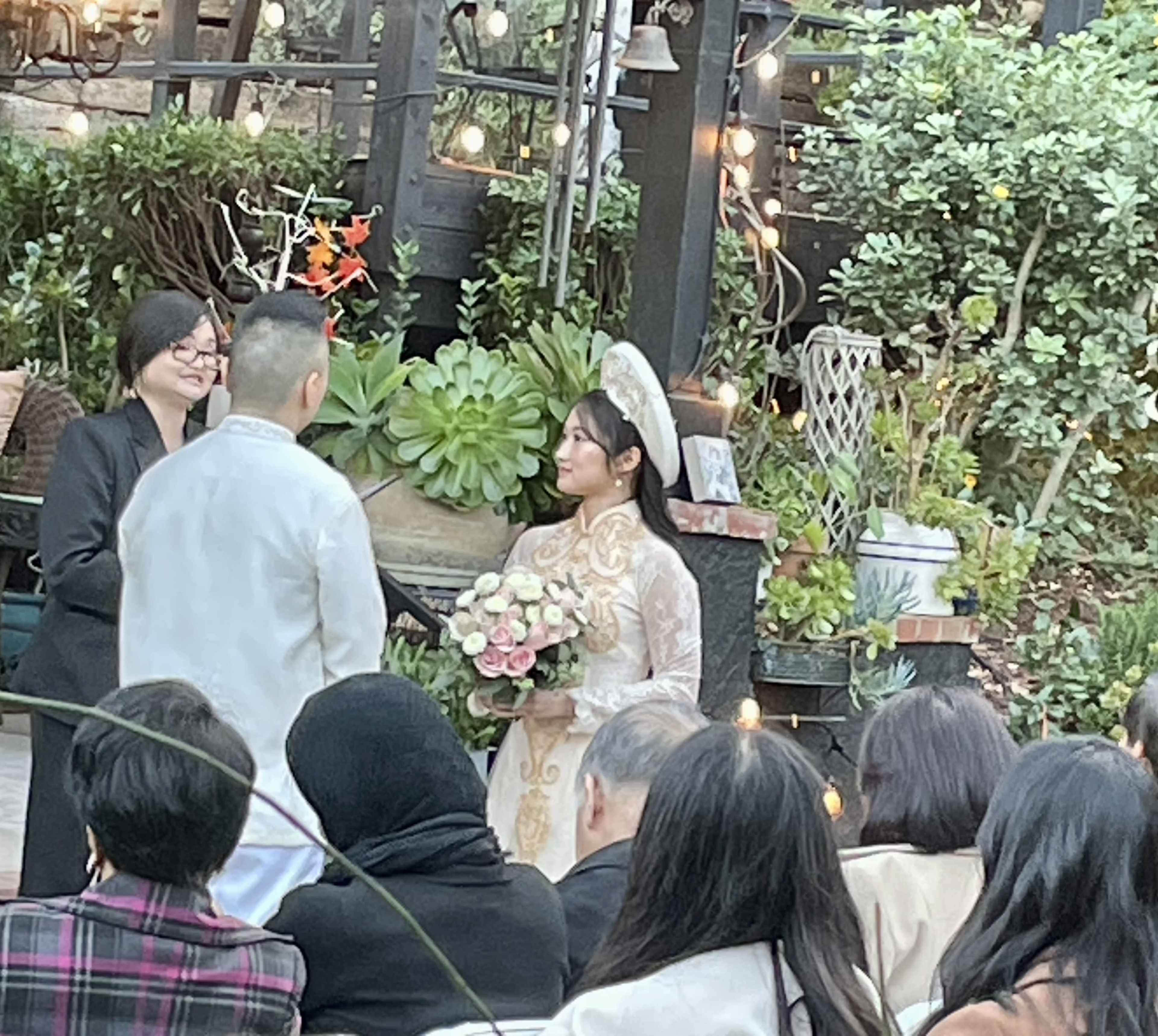 A bride in a traditional dress holds a bouquet while standing with a groom during a wedding ceremony outdoors, surrounded by greenery and string lights.
