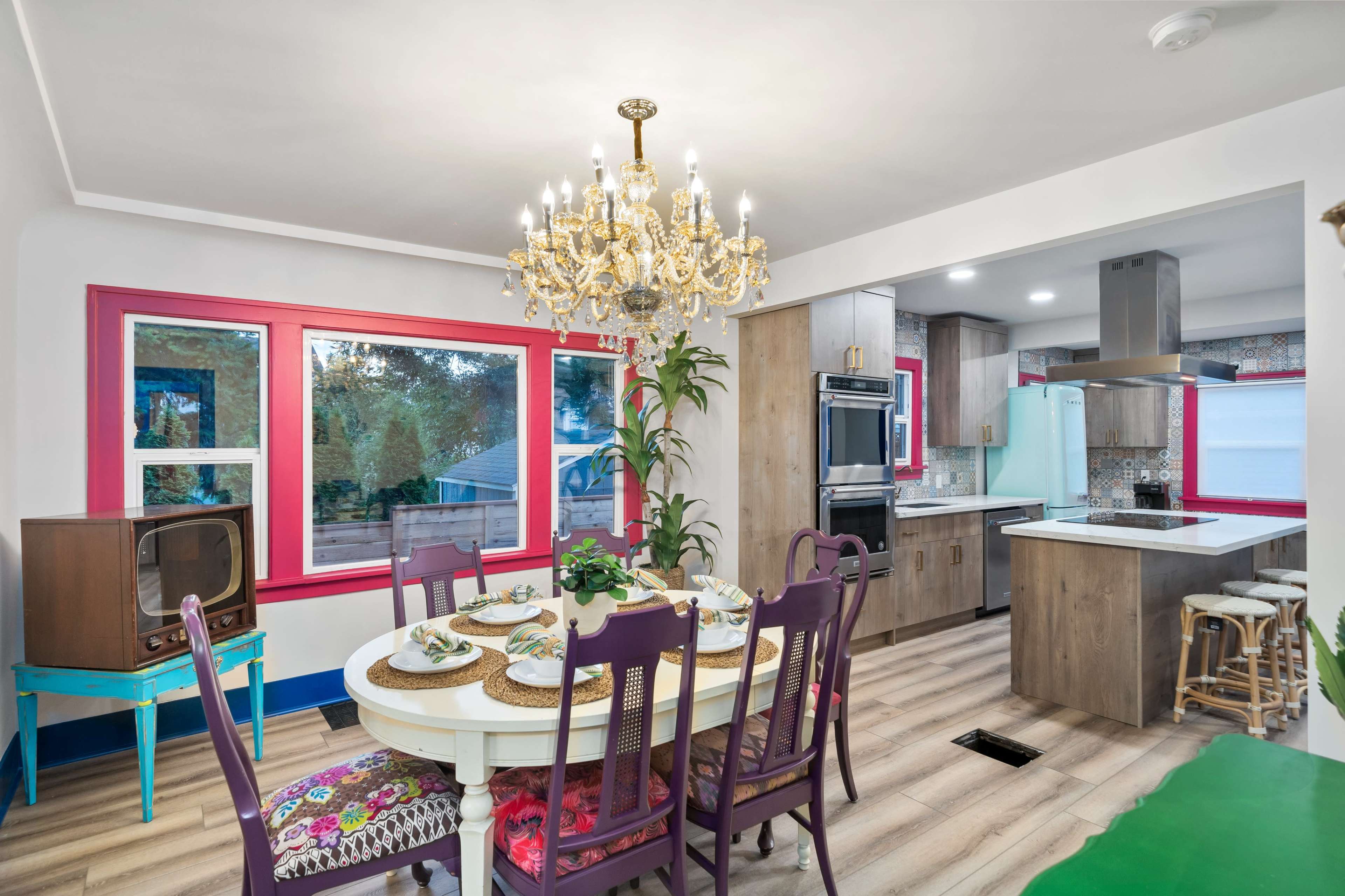 A dining area features a round table set for a meal, surrounded by colorful chairs, with a chandelier overhead and a view of a kitchen in the background.
