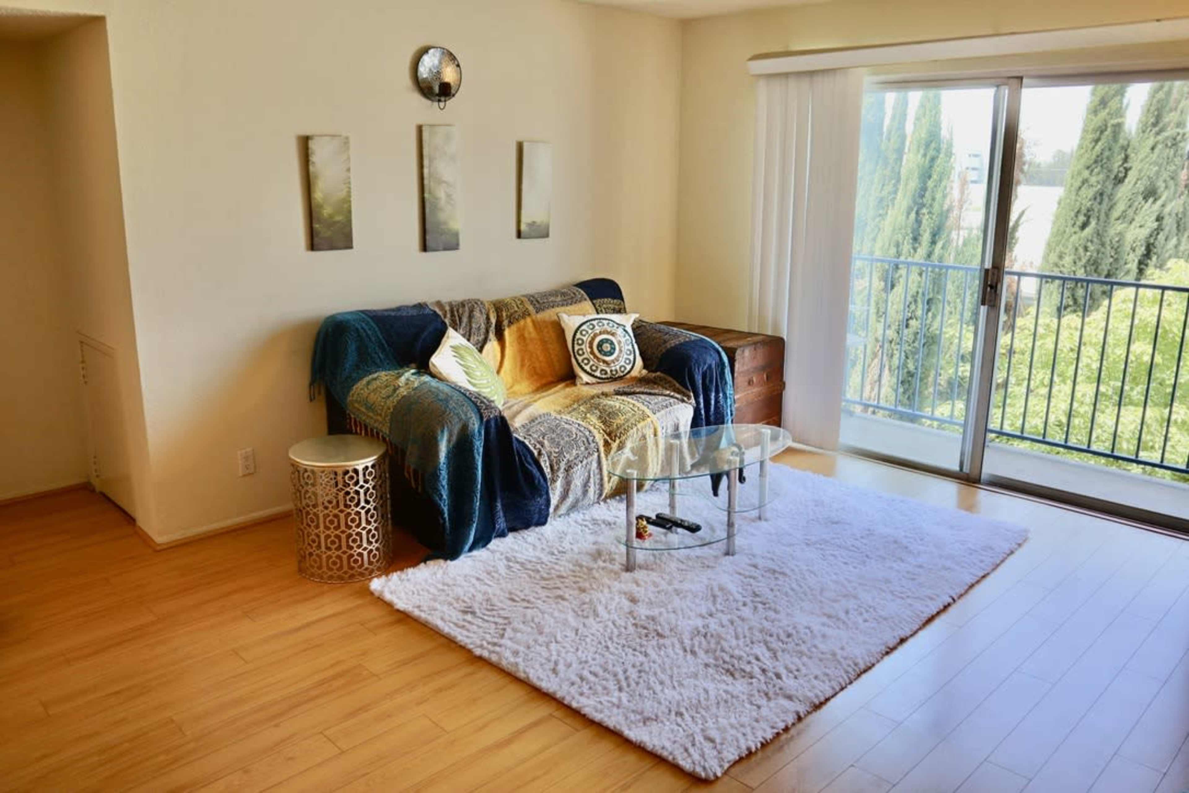 A cozy living room with a sofa draped in blankets, a glass coffee table, and a large rug on a wooden floor, featuring a view of trees through sliding glass doors.