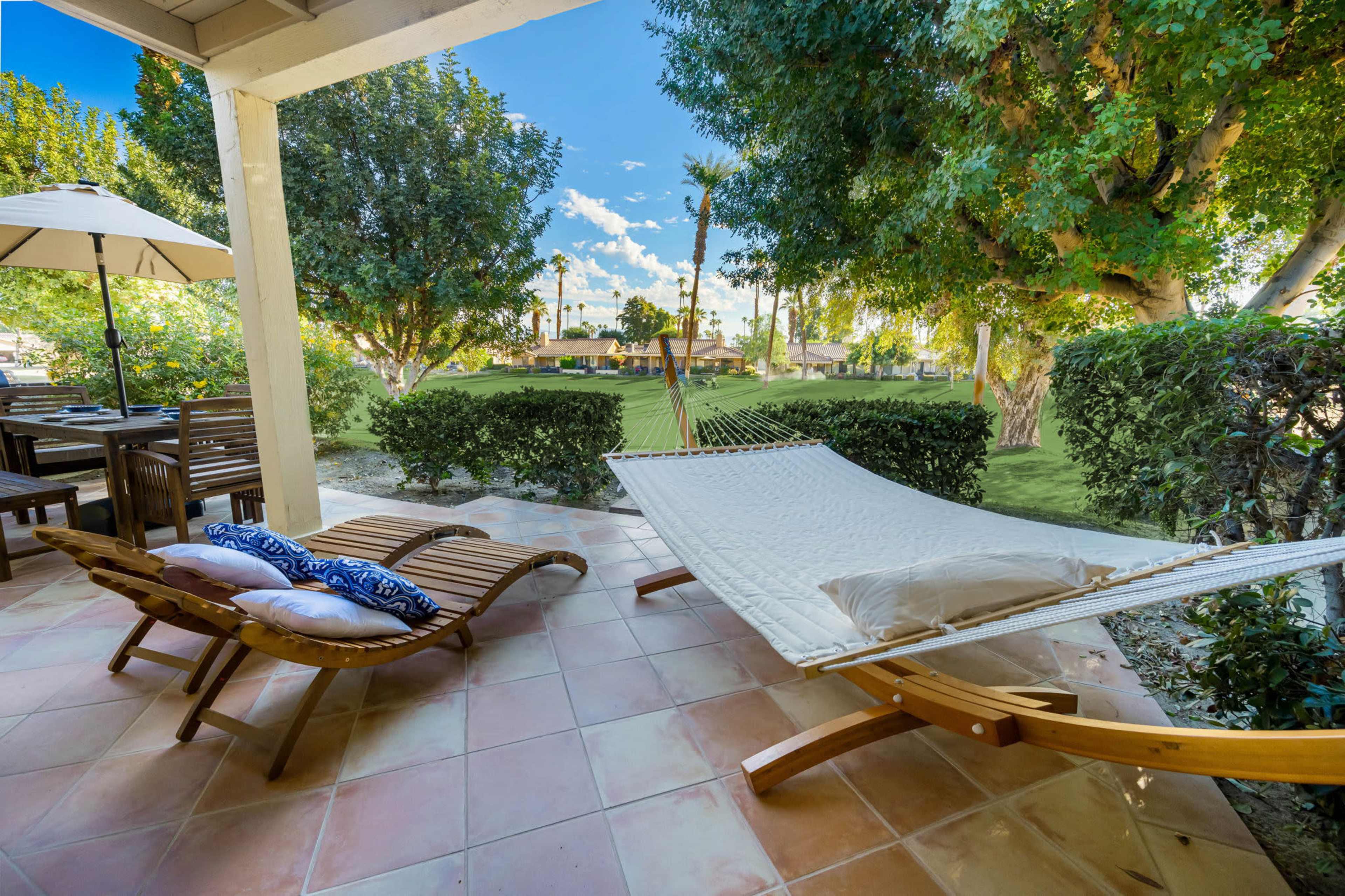 A hammock and a lounge chair are positioned on a patio overlooking a green lawn and trees.