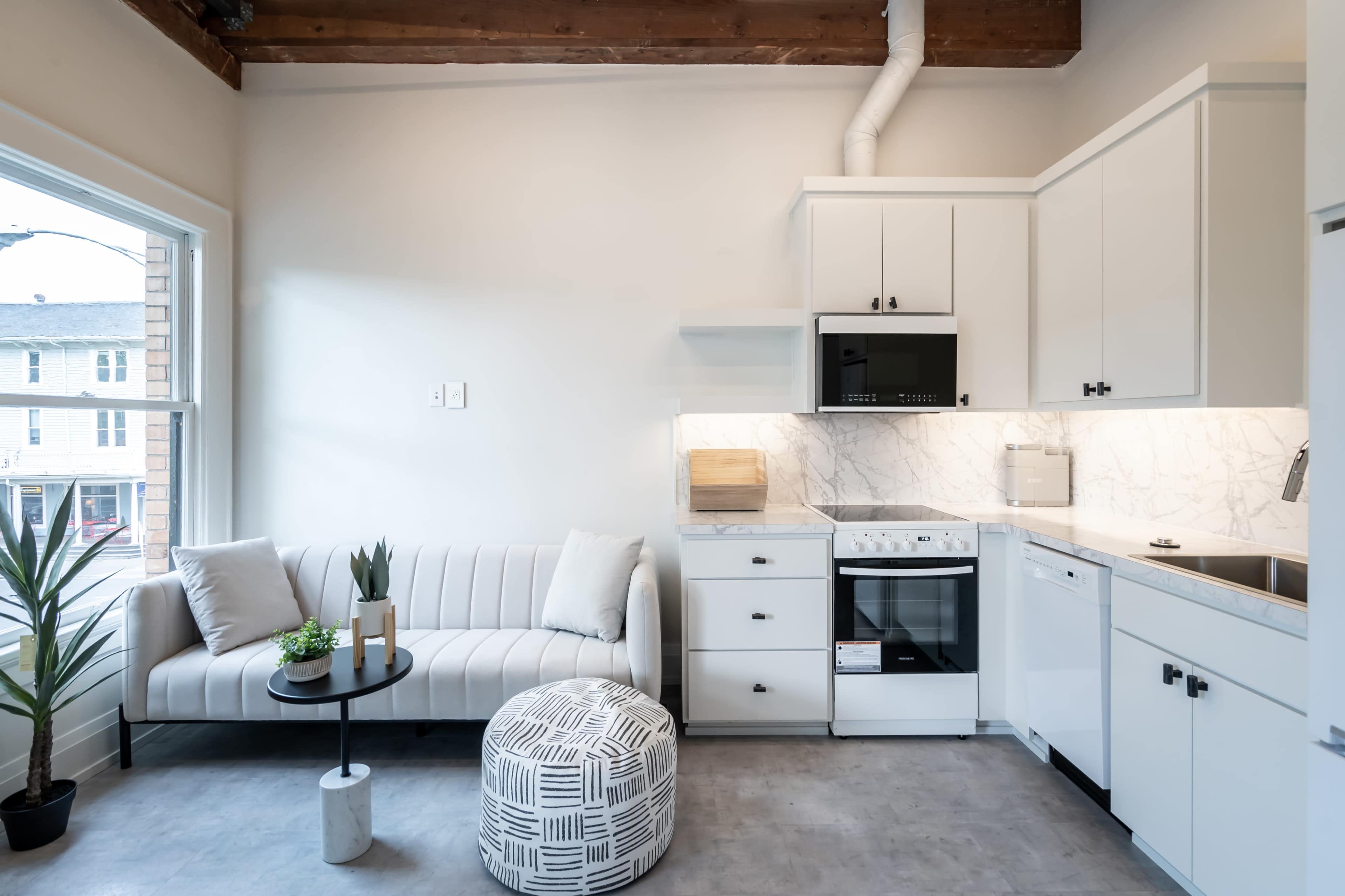 A modern kitchen and living area with a white sofa, a round coffee table, and a compact kitchen featuring light-colored cabinetry and a marble backsplash.