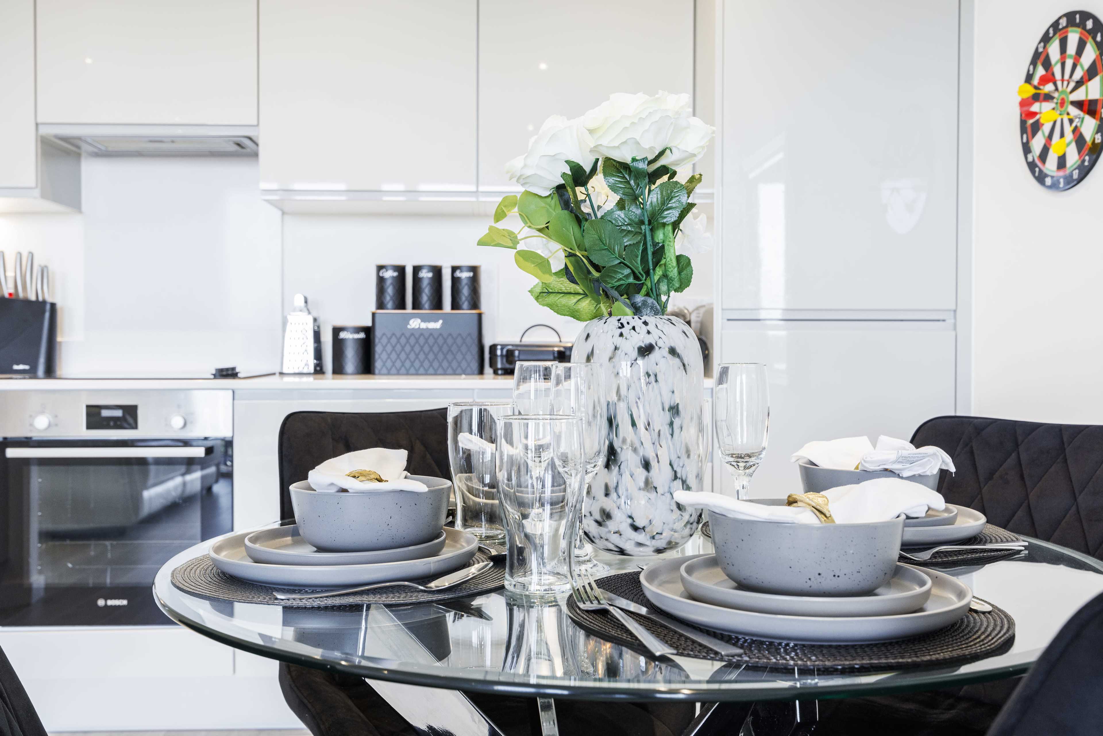 A dining table is set with gray plates and glassware, featuring a vase of flowers, in a modern kitchen with black appliances and a dartboard on the wall.