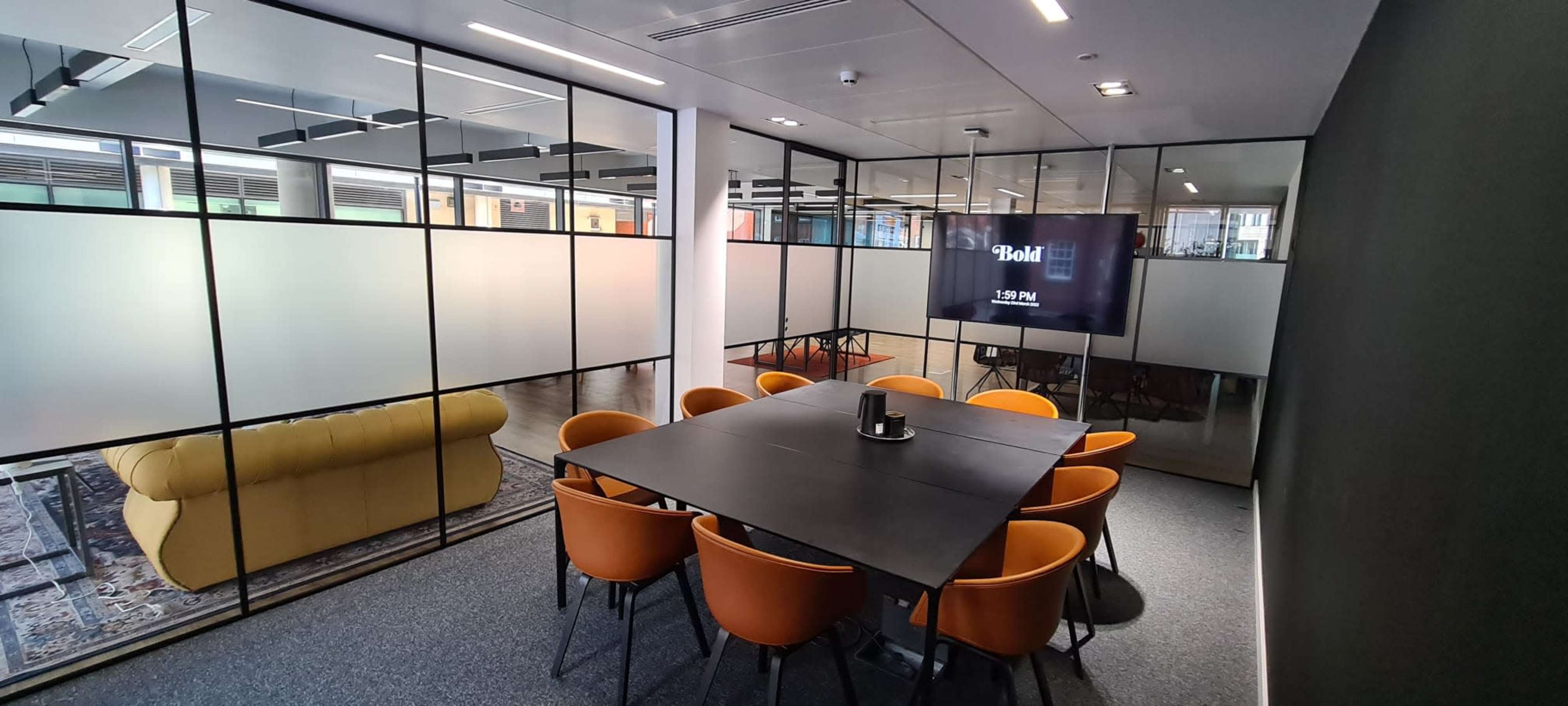 A modern meeting room with a black table surrounded by orange chairs, large glass walls, and a digital clock displaying the time.