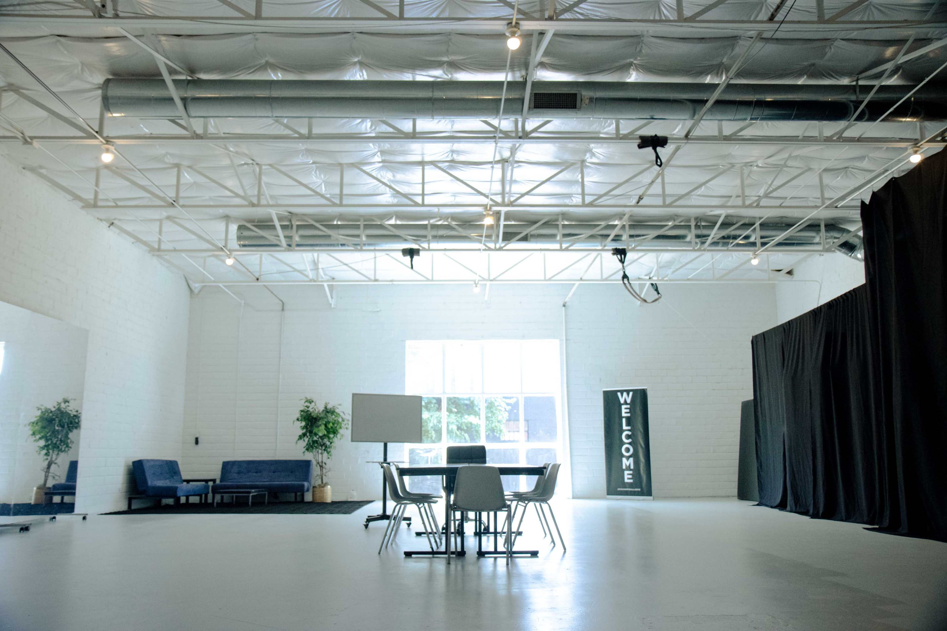 An empty, well-lit room with a white ceiling and walls, featuring a table with chairs, a sofa area, and a large "WELCOME" sign.