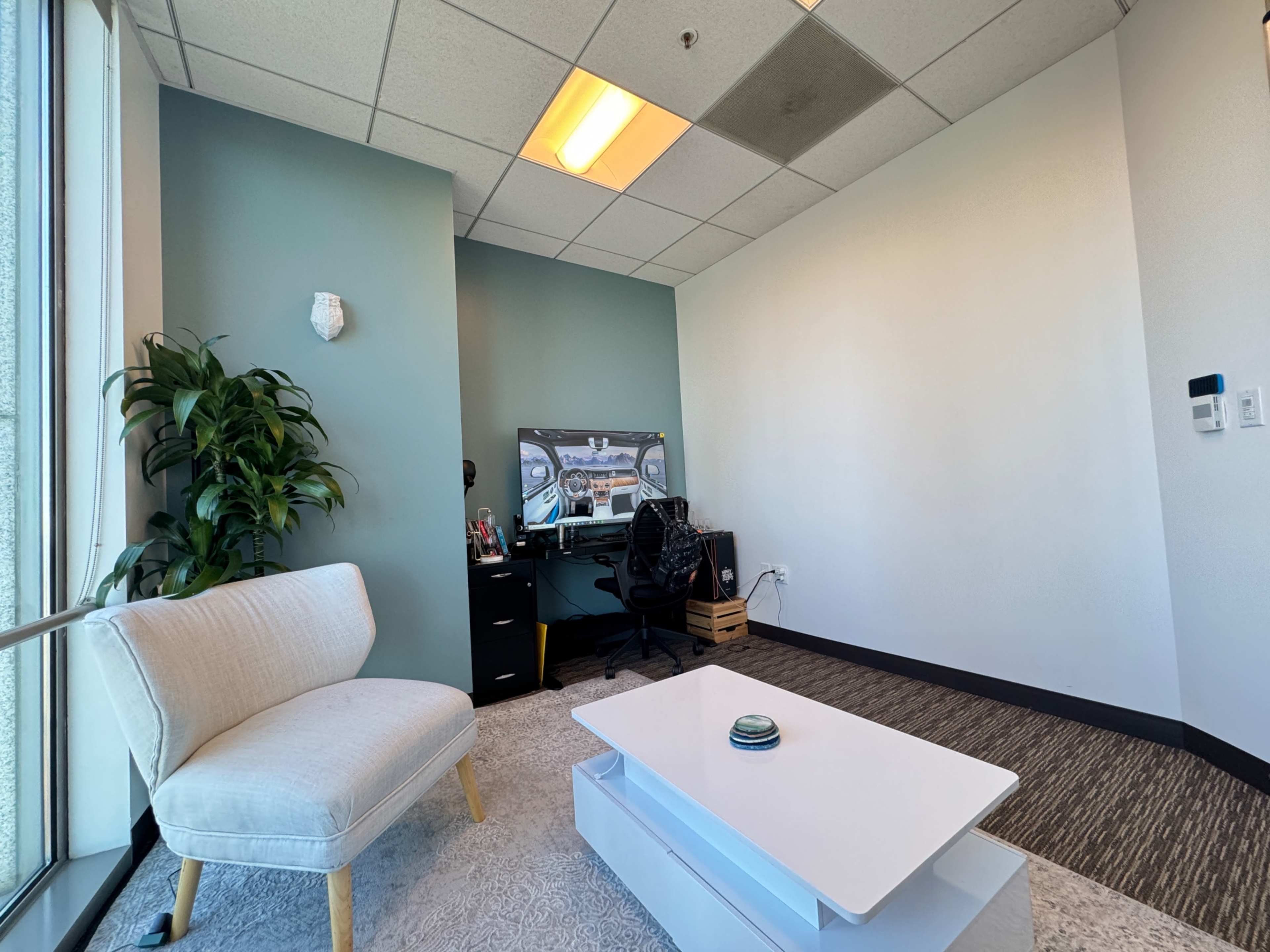 The image shows a modern office space with a white couch, a television, a desk equipped with a chair, and a potted plant.
