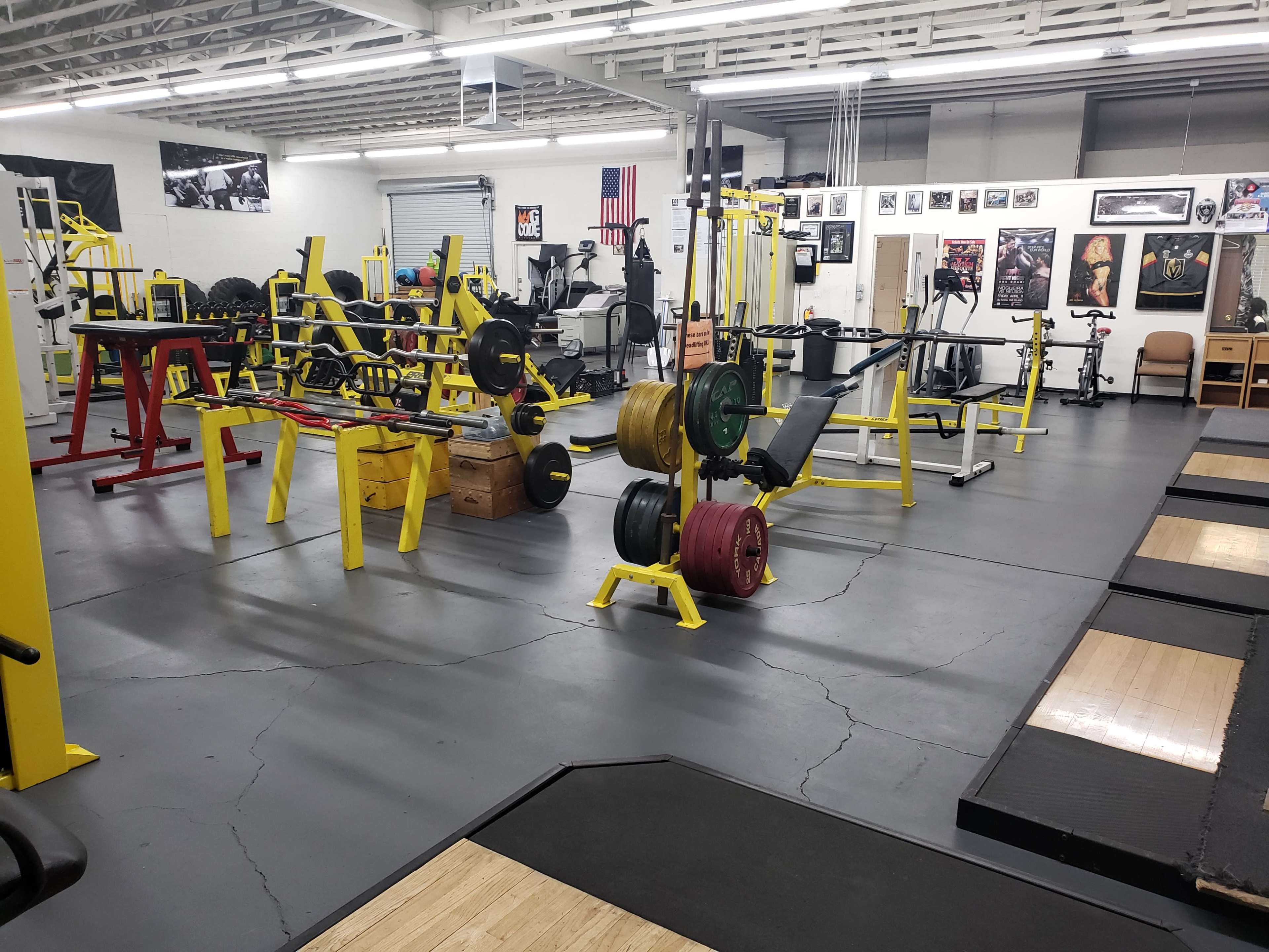 The image shows a gym filled with various weightlifting equipment, including benches, free weights, and machines, set against a backdrop of motivational images and flags.