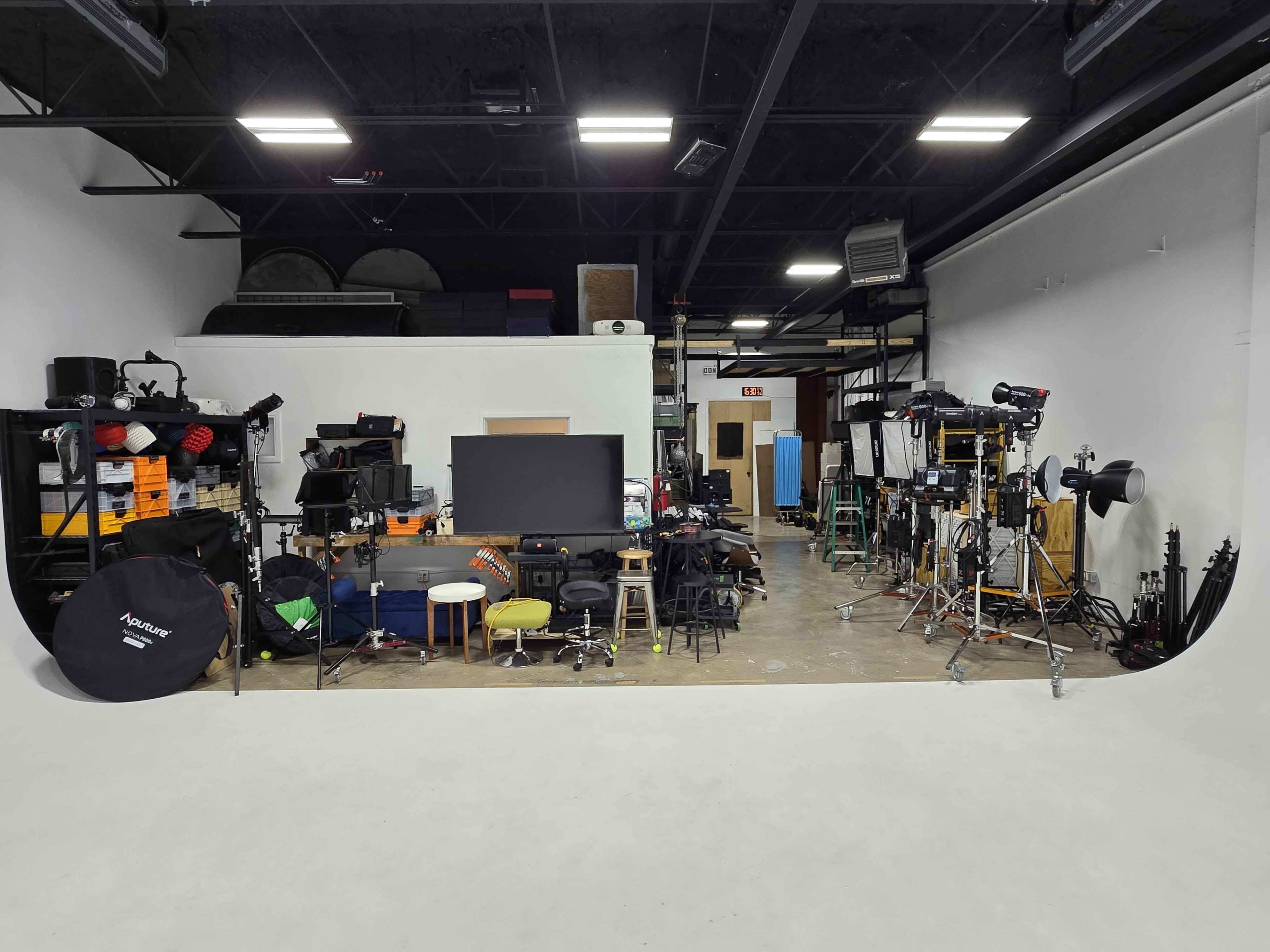 The image shows a photography studio filled with various equipment, including lighting, backdrops, and furniture, set against a plain wall.