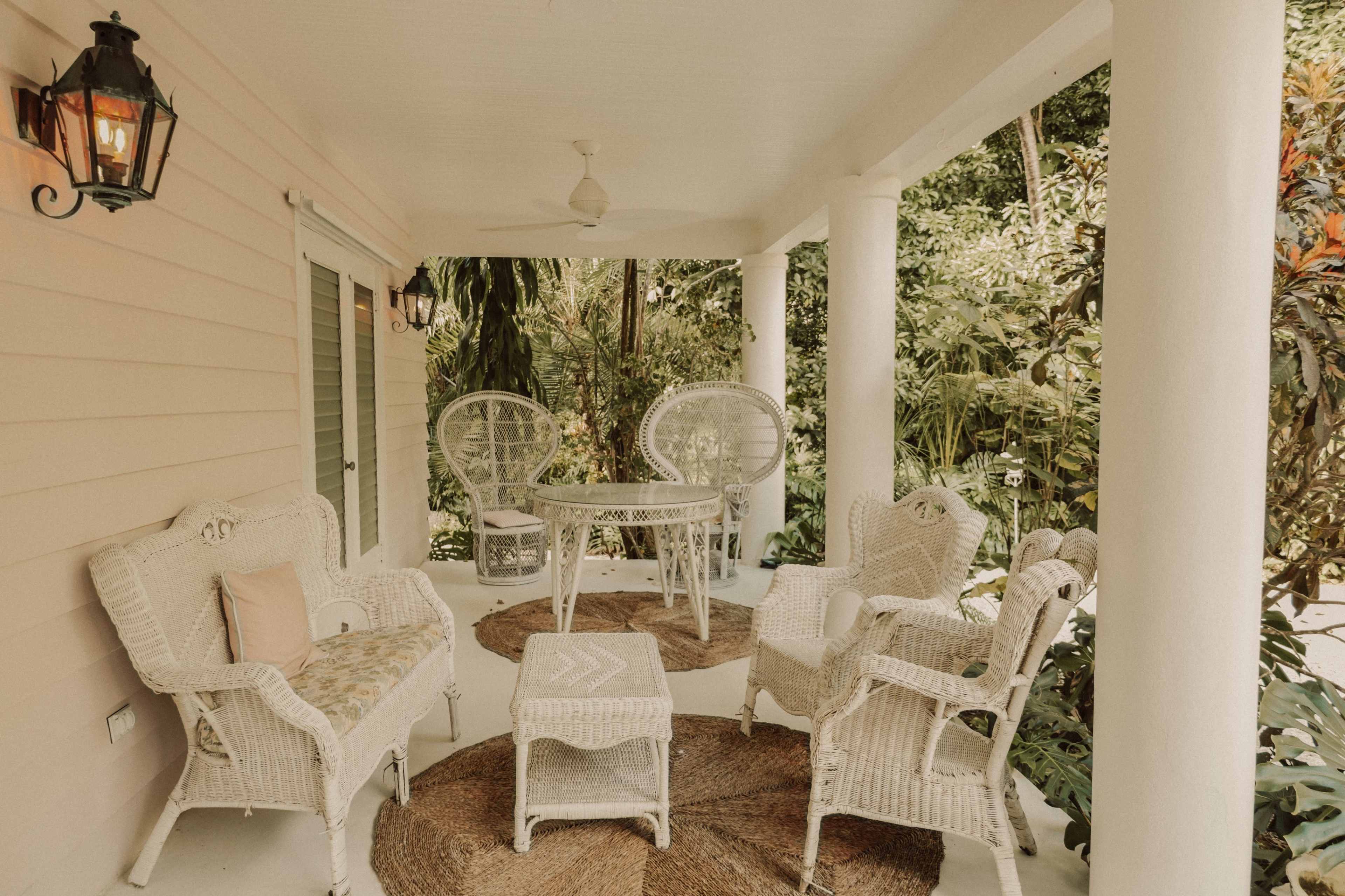 A covered porch features wicker furniture, including chairs and a table, surrounded by tropical greenery.