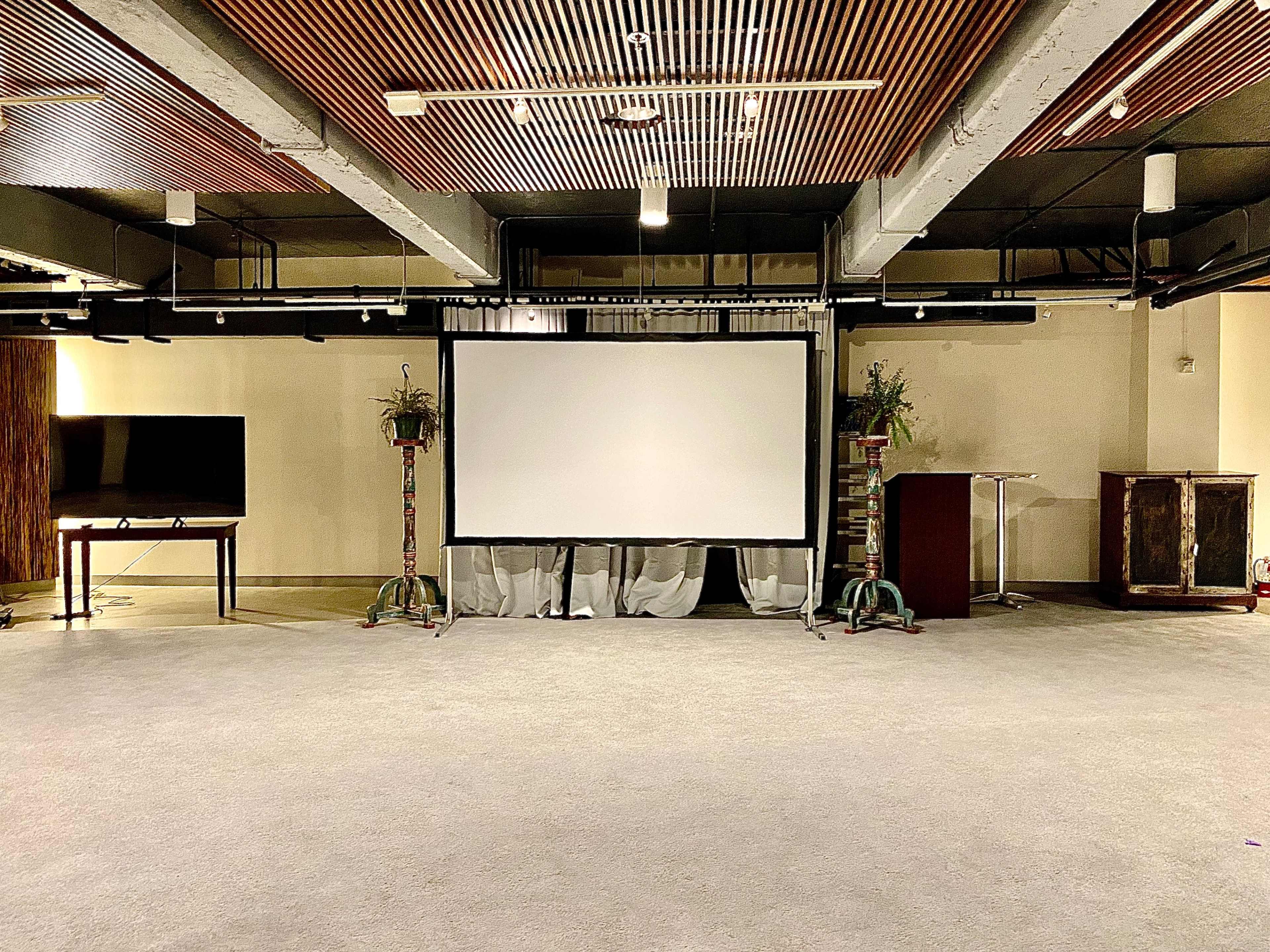 A spacious indoor venue featuring a large projection screen at the center, flanked by decorative plants and furniture.