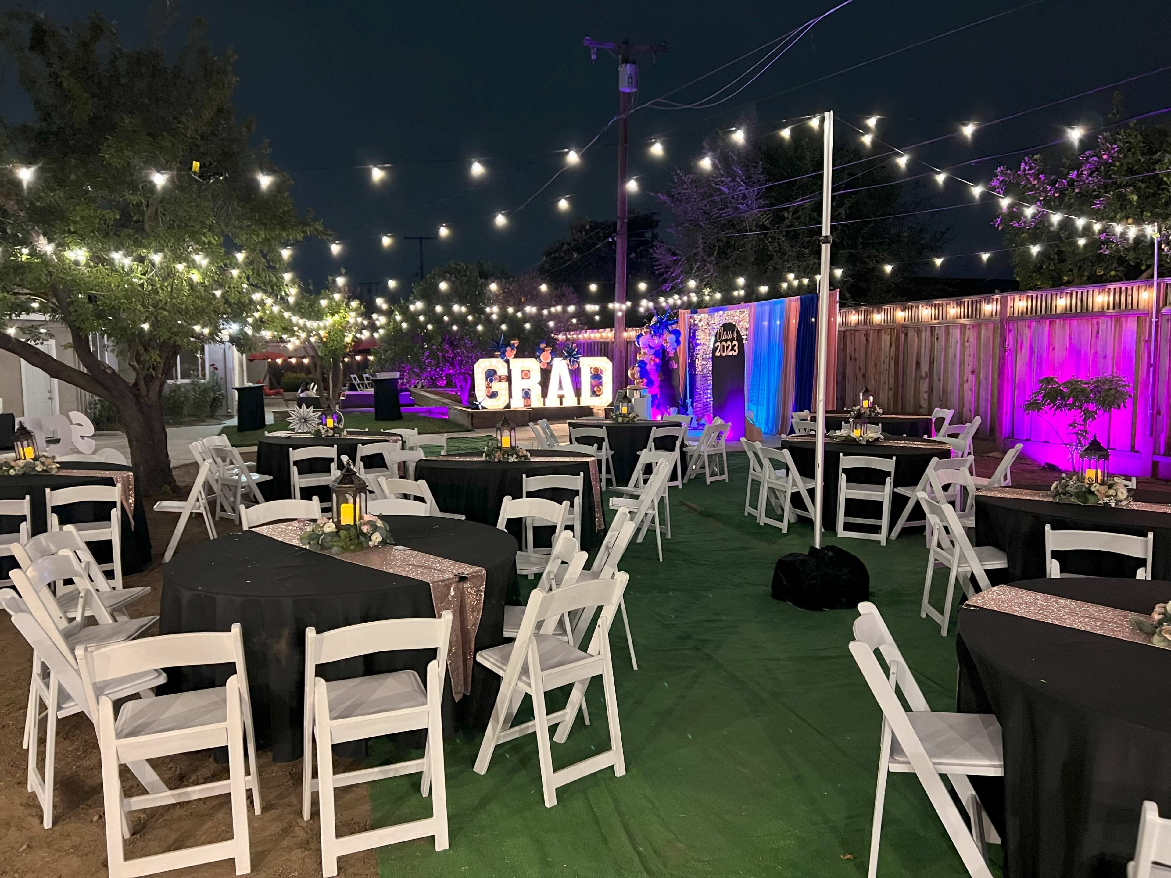 The outdoor event space is set up for a graduation celebration, featuring tables with white chairs and string lights illuminating the area.